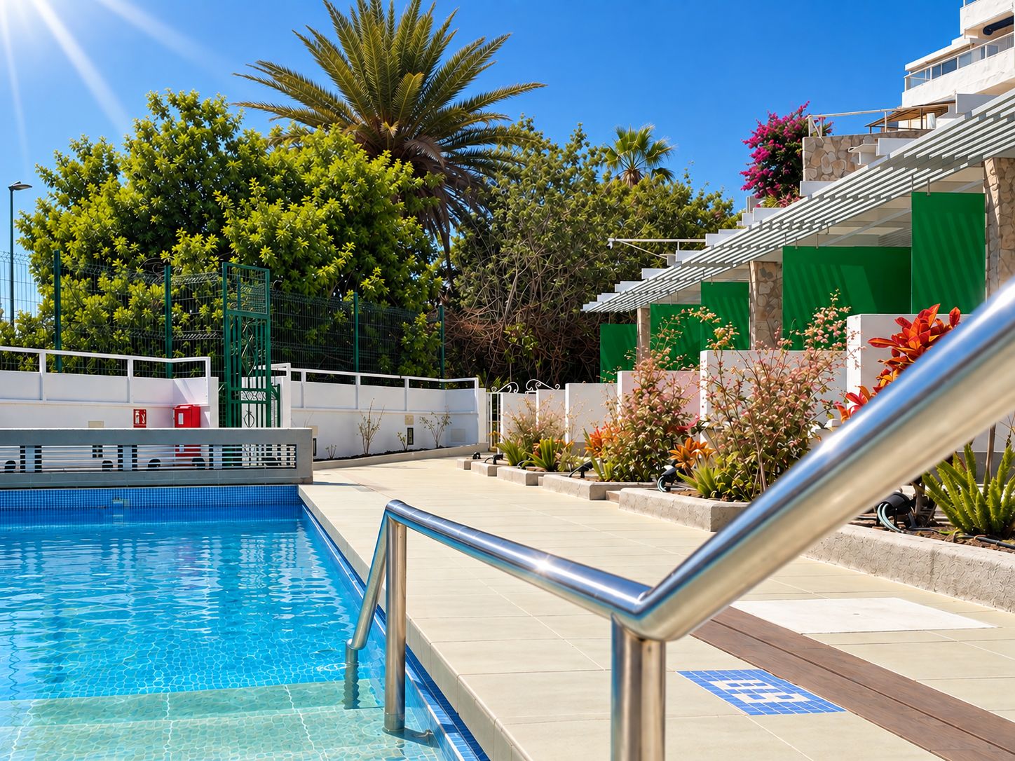 A swimming pool with a metal railing, surrounded by greenery and a building with a stone facade.