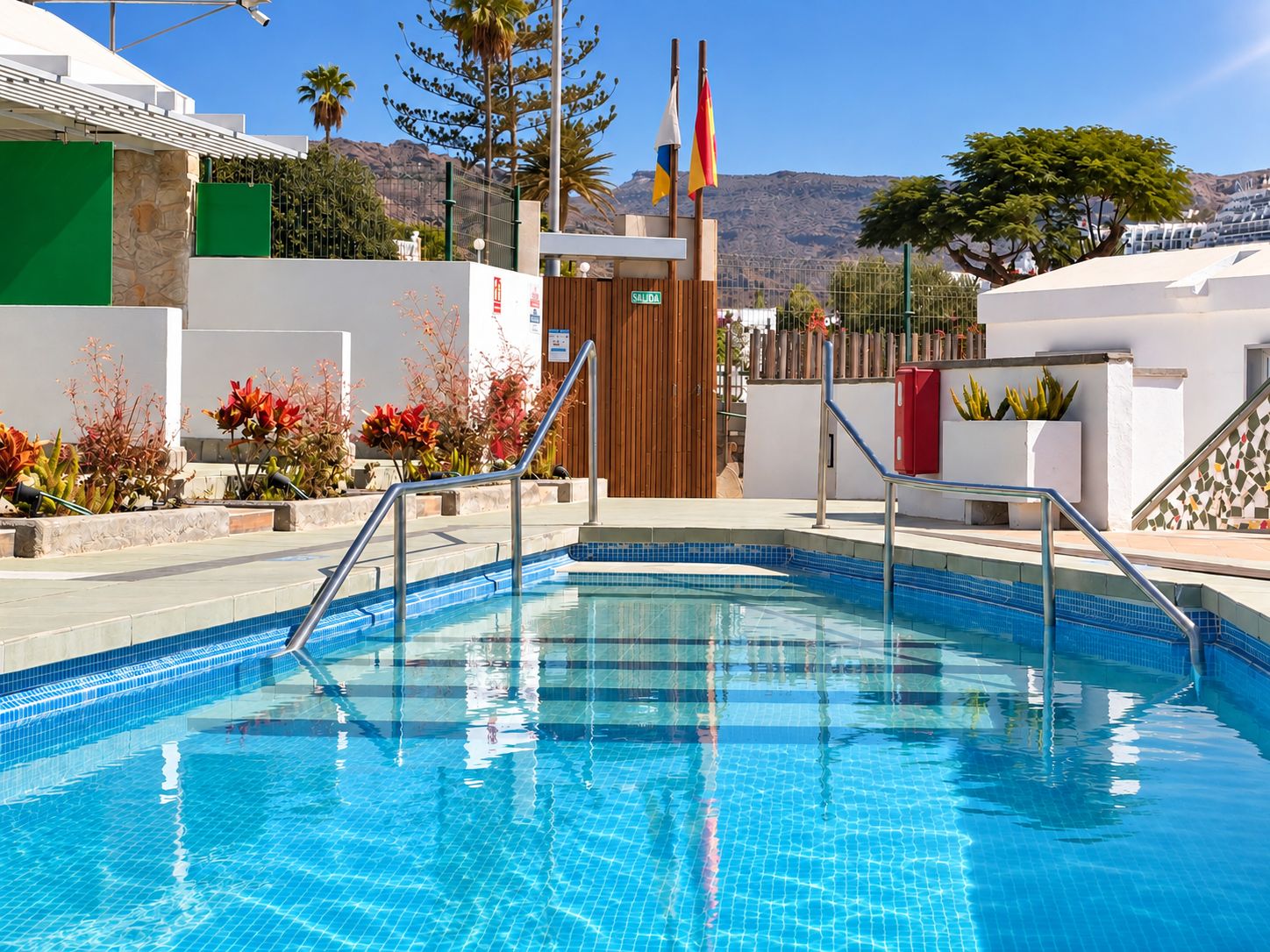 A swimming pool with a clear blue tile design, surrounded by a white building with a wooden door and a sign that reads 'SALIDA'.