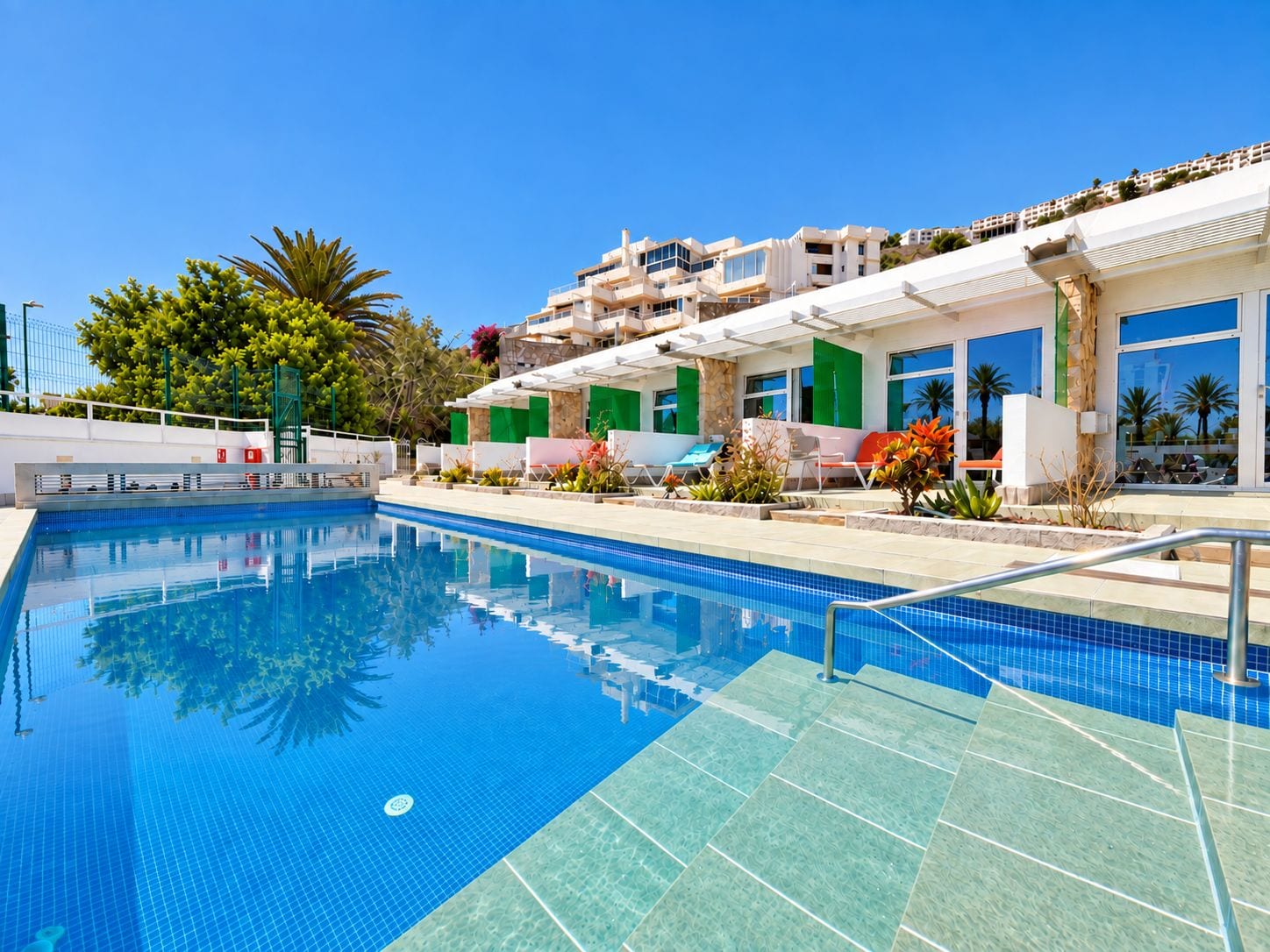 A swimming pool with clear blue water, surrounded by a poolside area with lounge chairs and potted plants.