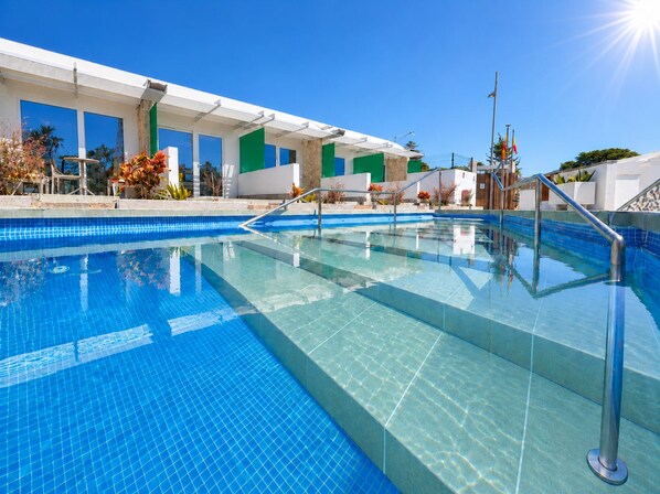 Une piscine d’un bleu éclatant, bordée d’une terrasse carrelée, entourée d’un bâtiment doté de grandes fenêtres et d’un mur d’accent vert.