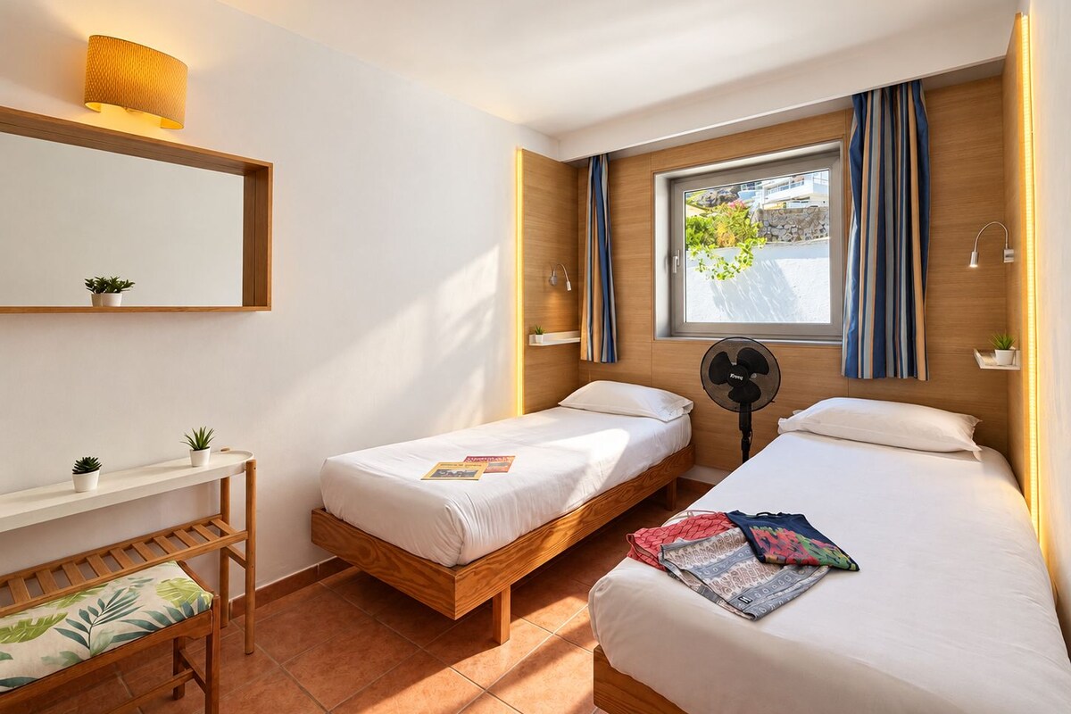 A hotel room with two single beds, a wooden bench, a small table with a plant, a window with curtains, and a wall-mounted fan.