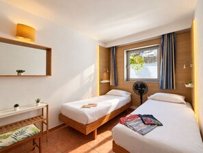 A hotel room with two single beds, a wooden bench, a small table with a plant, a window with curtains, and a wall-mounted fan. - Leticia Del Mar (Mogan)