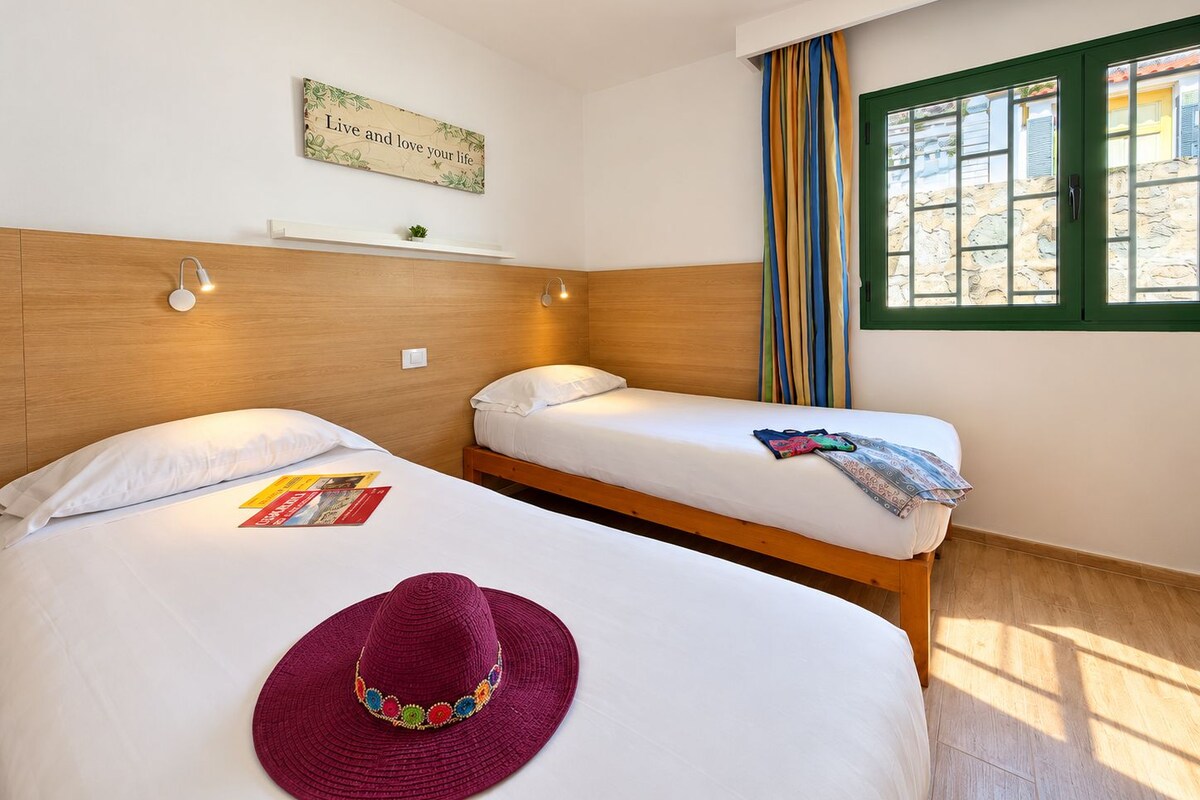 A hotel room with two beds, a wall-mounted shelf with a plant, a window with curtains, and a motivational quote on the wall.