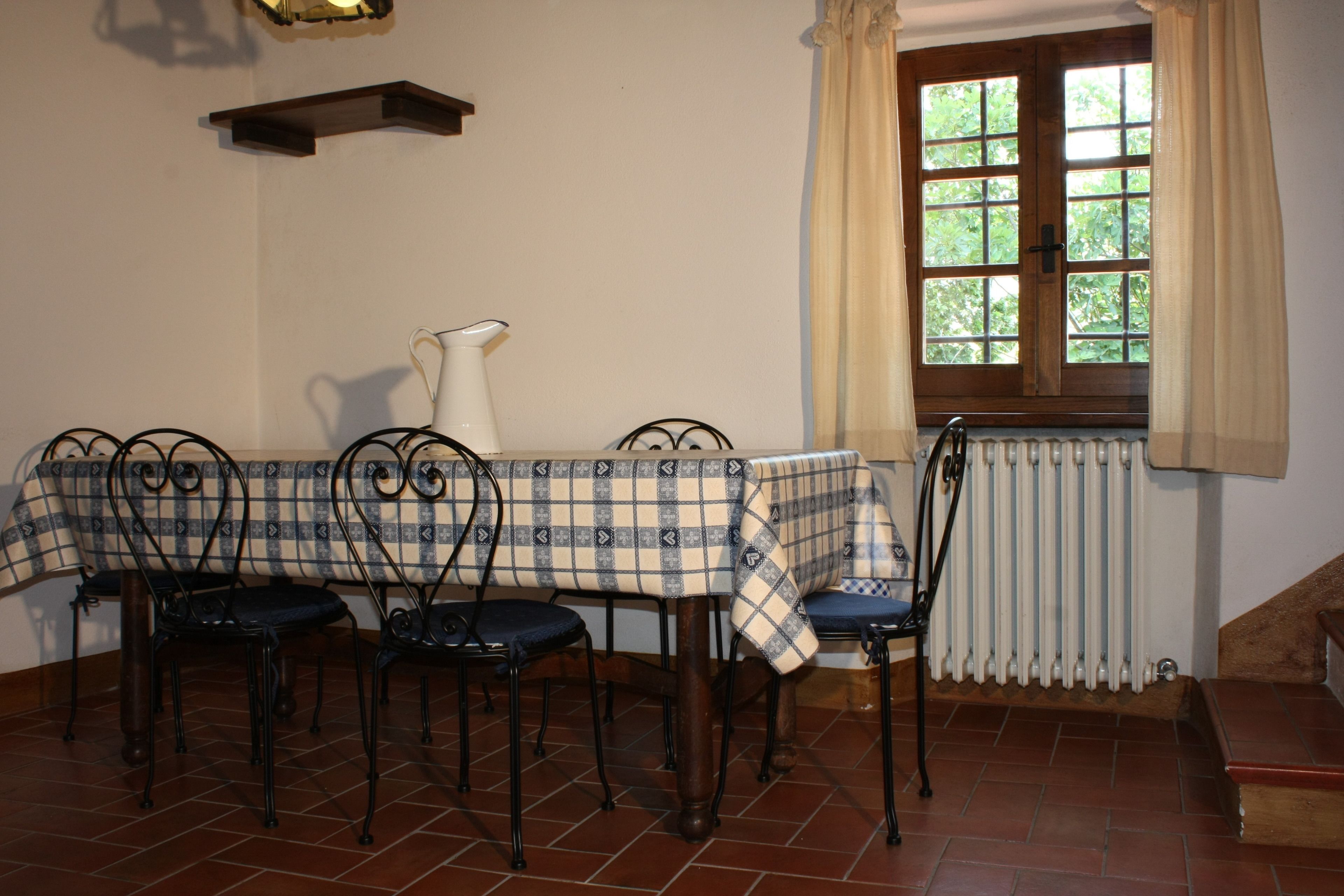 Un comedor con mantel a cuadros, cuatro sillas, una jarrón blanco, un radiador y una ventana con cortinas.