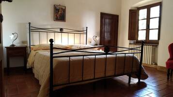 A bedroom with a metal bed frame, a bedside table, a lamp, a window with curtains, and a radiator.