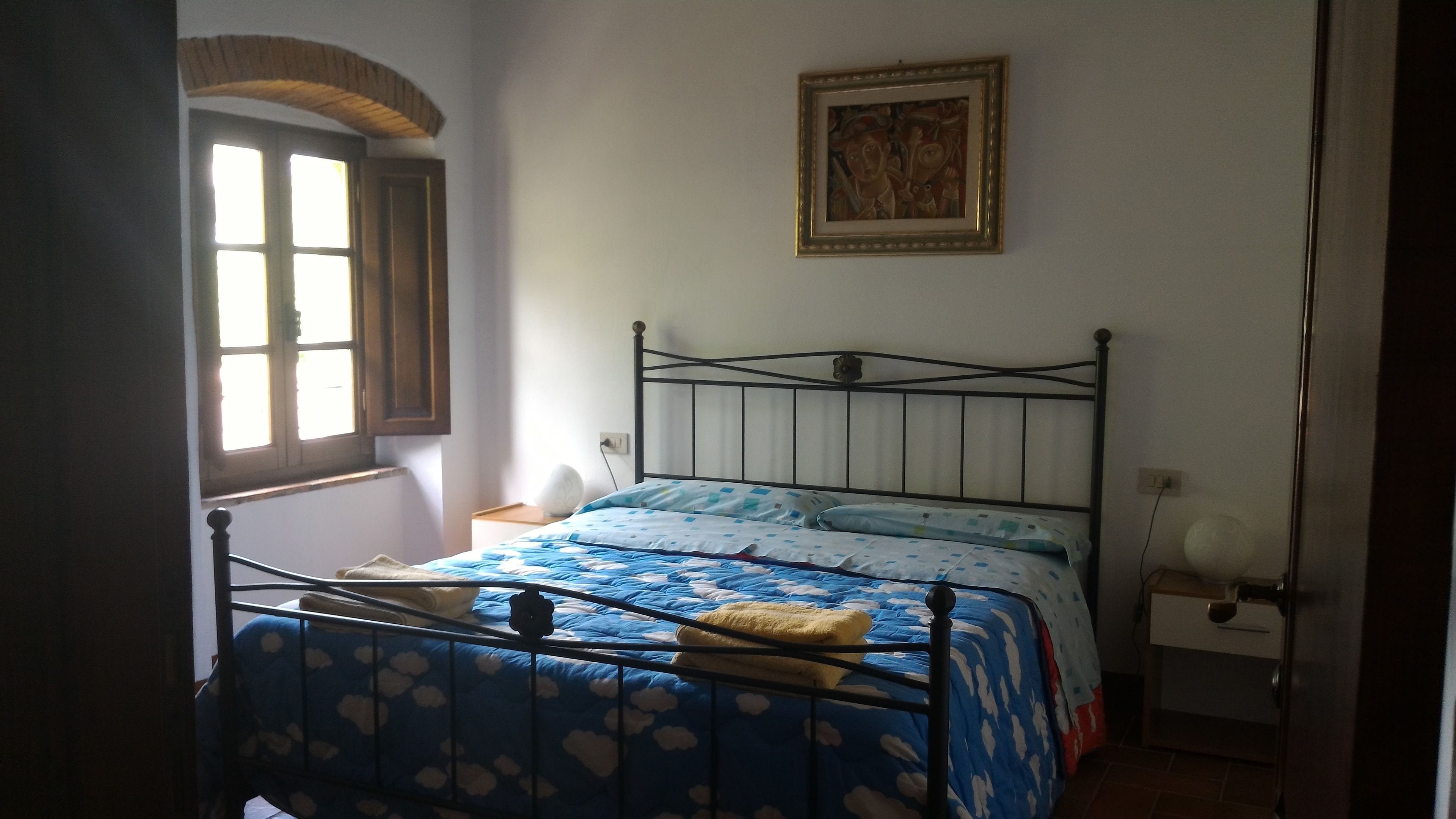 Een slaapkamer met een bed, een raam, een nachtkastje en een aan de muur hangend schilderij.