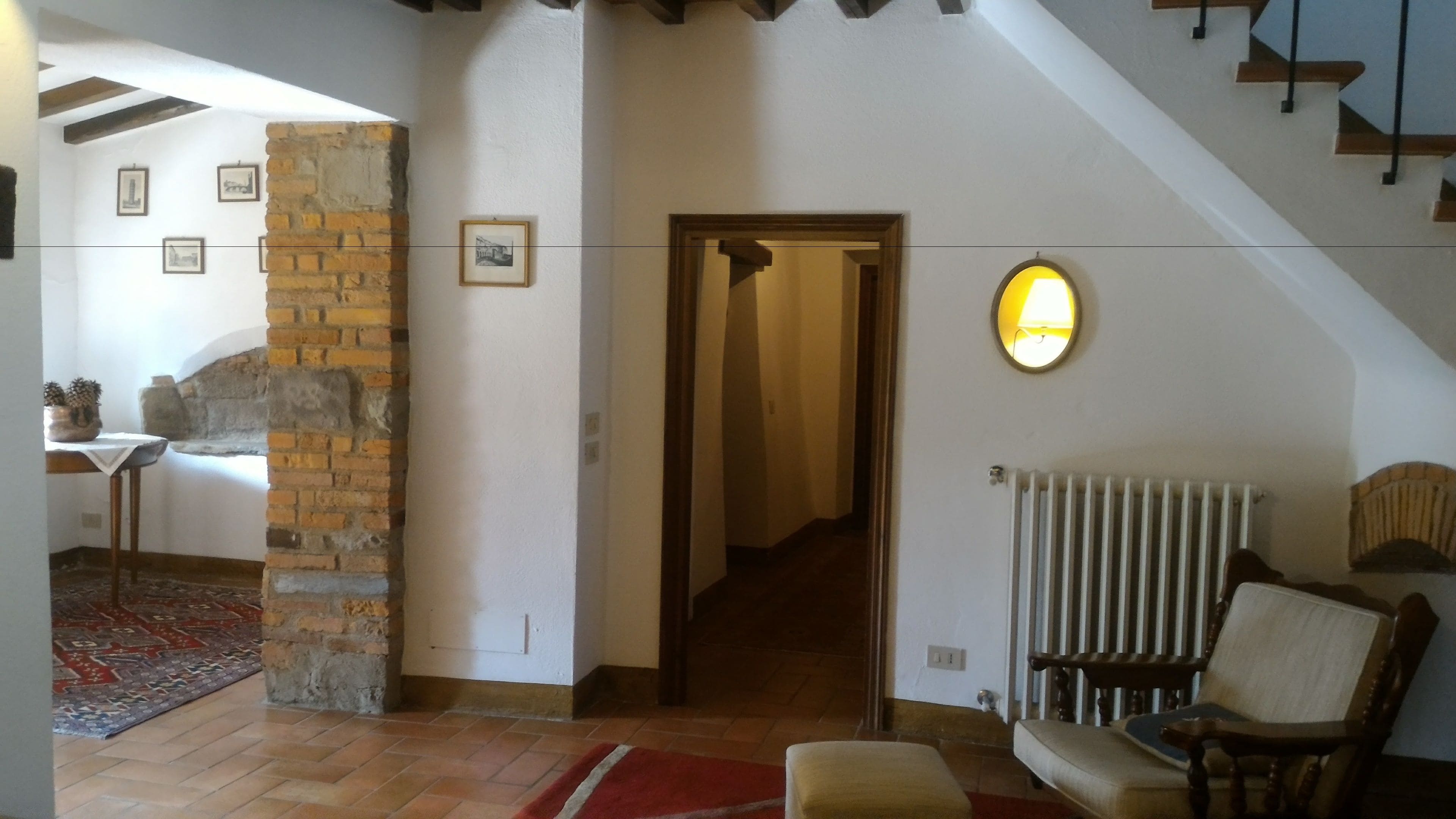 Una stanza spaziosa con soffitto alto, pareti in mattoni a vista, una scala in legno e un’accogliente area relax con poltrona e poggiapiedi.