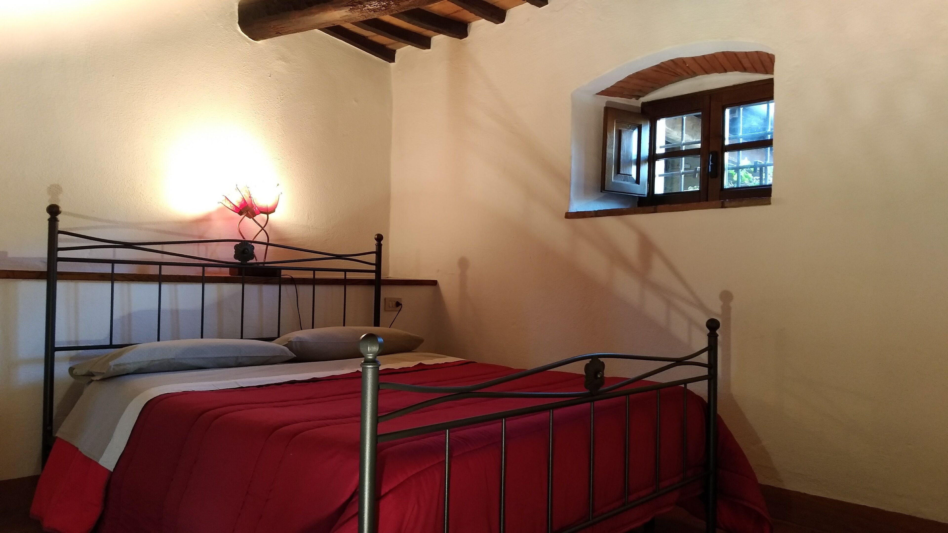 A single bed with a red bedspread, a nightstand with a lamp, and a small window with a view of greenery.