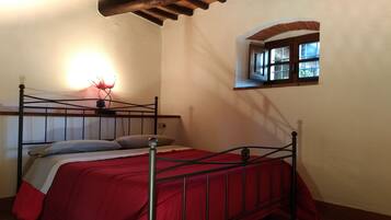 A single bed with a red bedspread, a nightstand with a lamp, and a small window with a view of greenery.