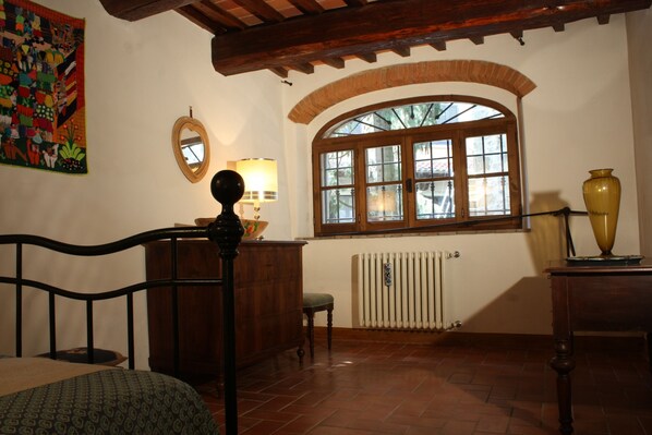 A room with a bed, a wooden desk, a chair, a window with an arched design, a radiator, and a wall with a mirror and a colorful painting. - B&B Villa La Nussa (Capolona)