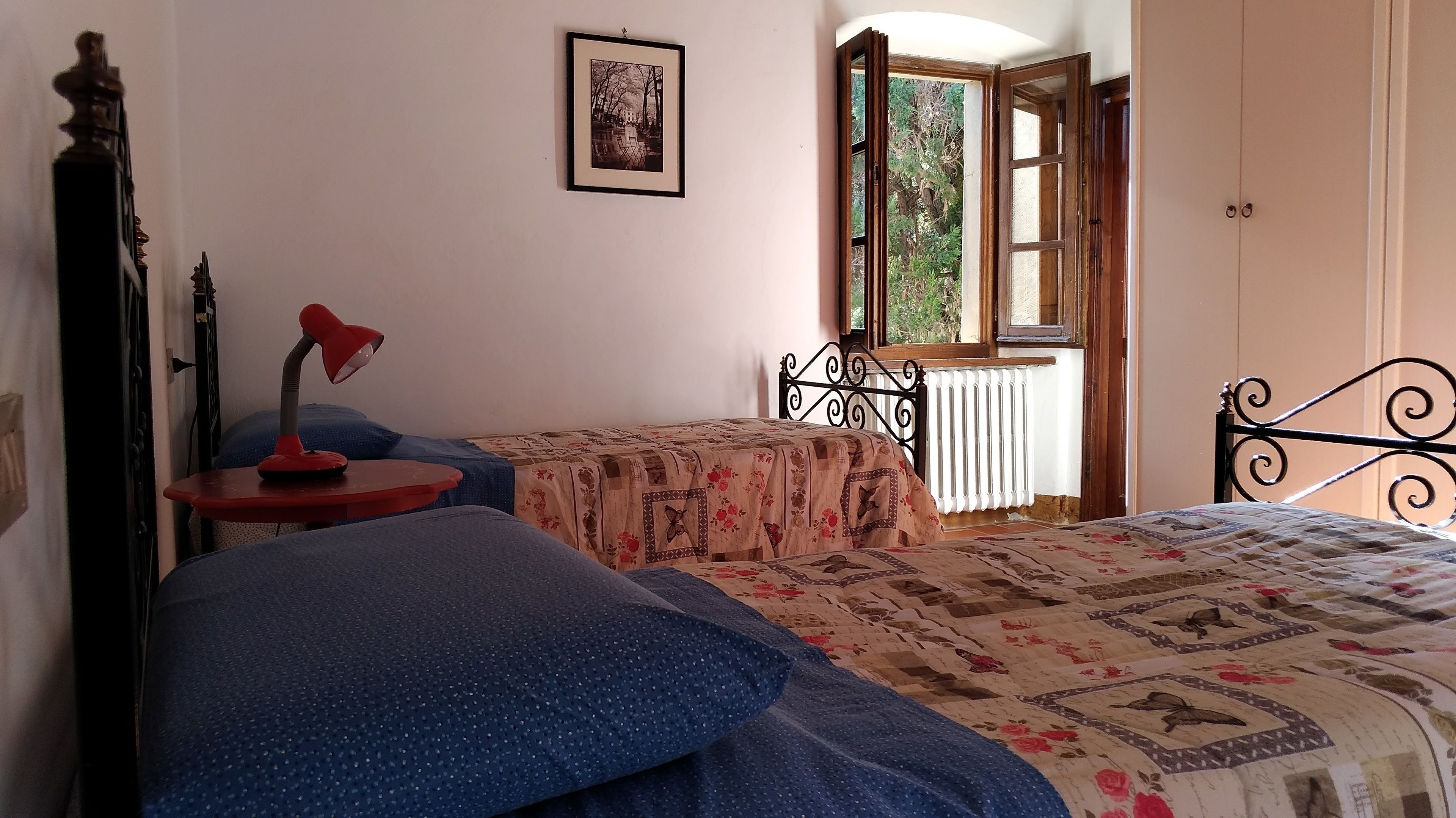 Un dormitorio con dos camas, una mesita con una lámpara, un radiador y una ventana con vistas a zonas verdes.