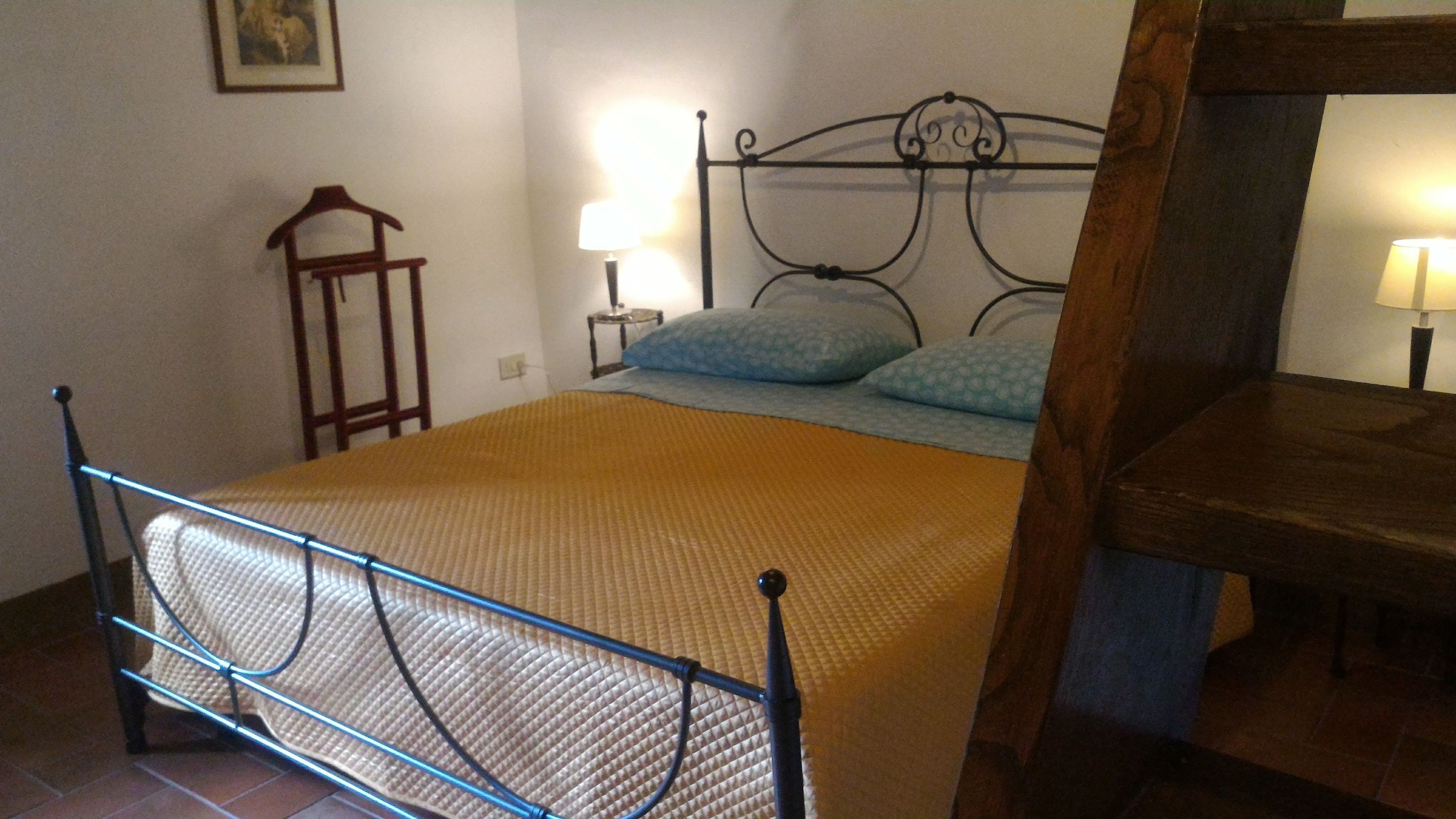 A bed with a metal frame, a wooden headboard, and a patterned bedspread. There is a nightstand with a lamp and a small table with a lamp.