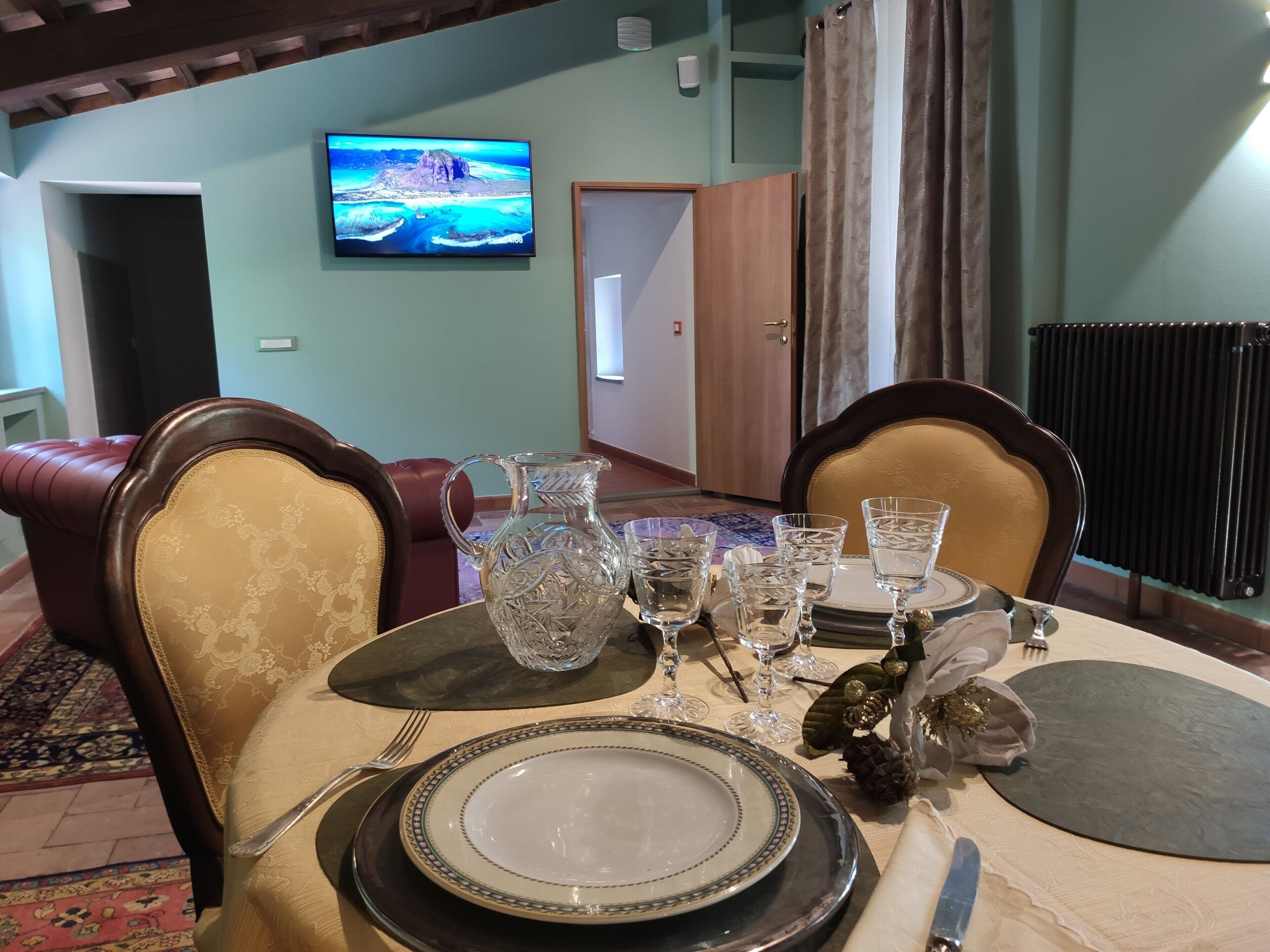 Una mesa de comedor preparada con platos, vasos y una jarrón, con un televisor que muestra un paisaje pintoresco al fondo.