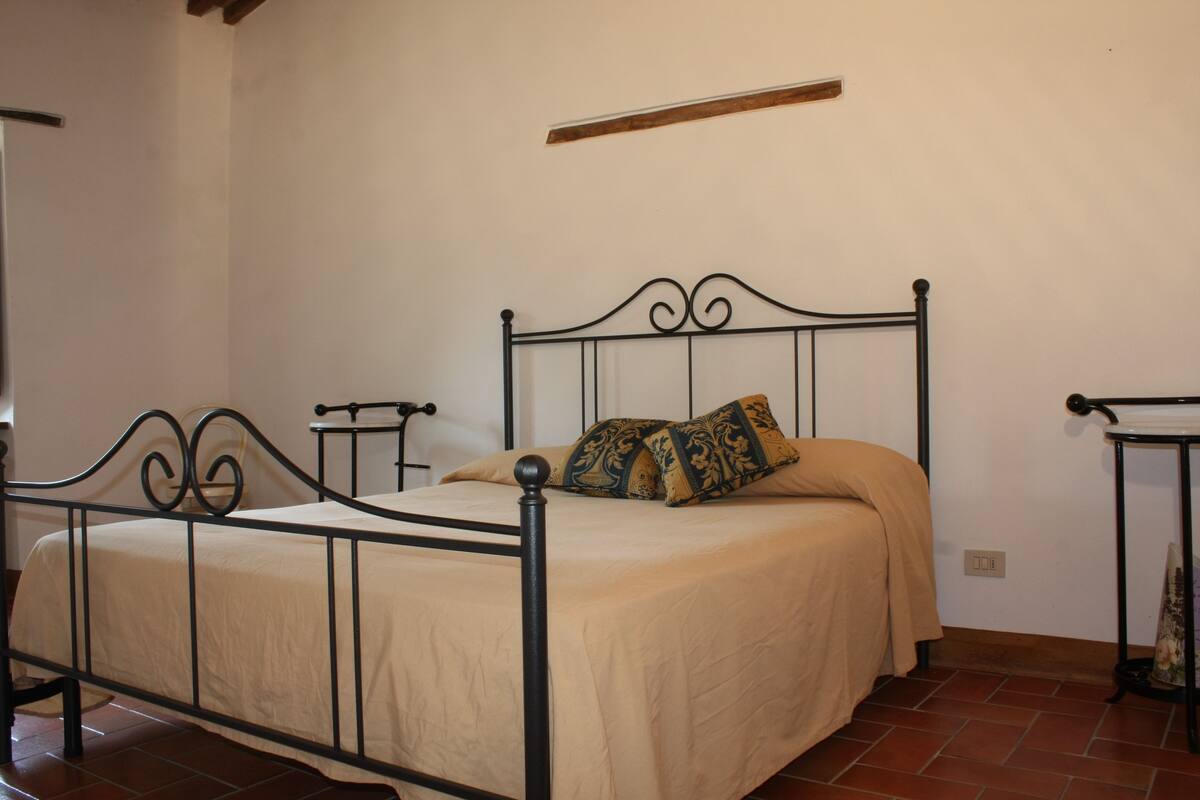 A single bed with a metal frame, a beige bedspread, and a decorative pillow. There is a small bedside table with a lamp on it. The room has white walls and a tiled floor.