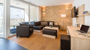 A modern living room with a sectional sofa, a coffee table, a desk with a laptop, and a large window offering a view of the outside. - Centerpark Apartments (Visp)
