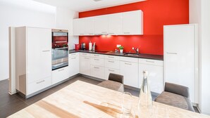 A modern kitchen with white cabinets, a red accent wall, and a wooden dining table with chairs. - Centerpark Apartments (Visp)