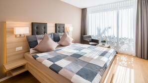 A modern hotel room with a large bed, two bedside lamps, a small table with a plant, and a view of the outdoors through sheer curtains. - Centerpark Apartments (Visp)