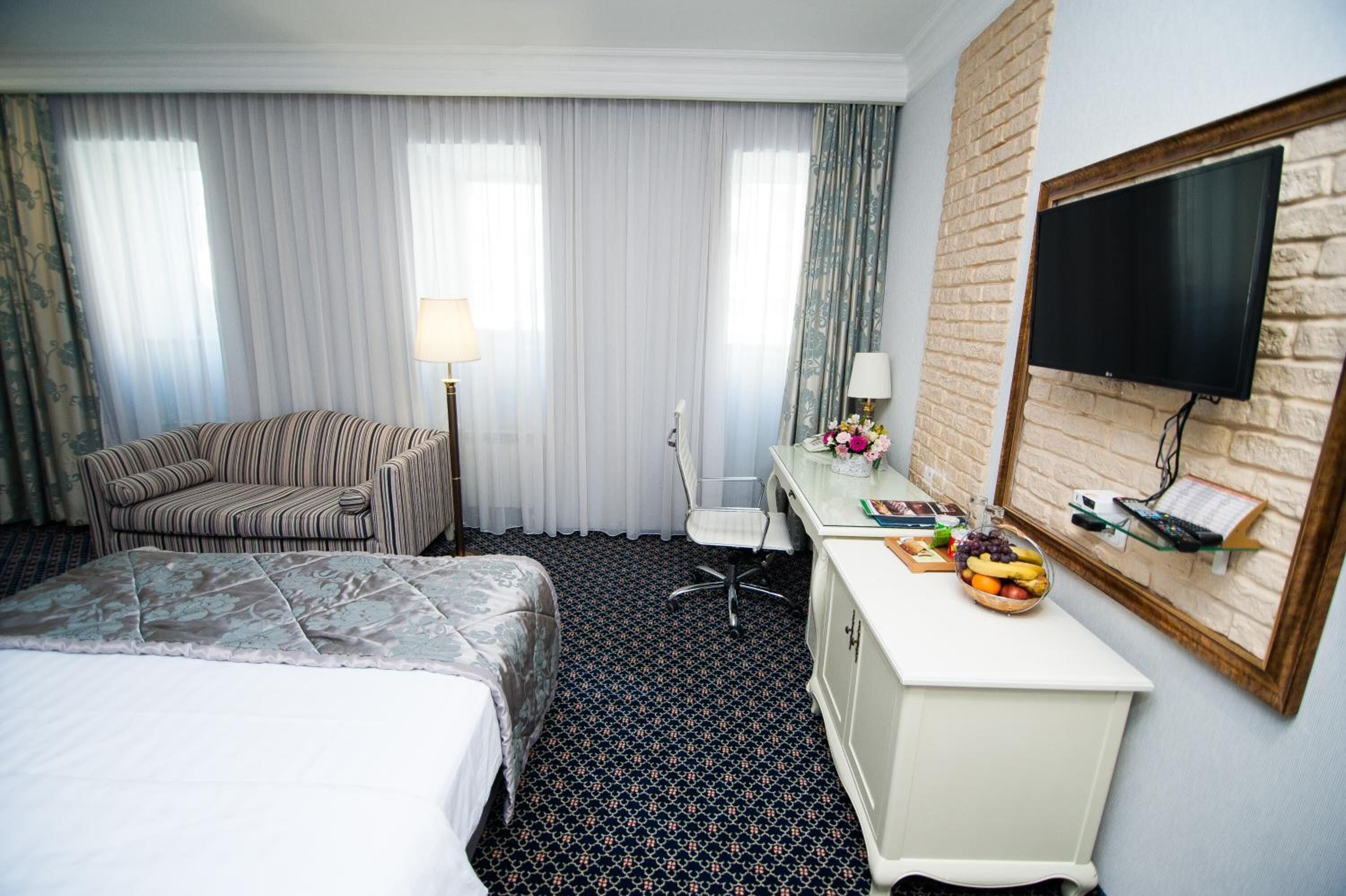 Habitación de hotel con una cama, un escritorio con una computadora y un frutero, una silla y un televisor fijado a una pared de ladrillo.