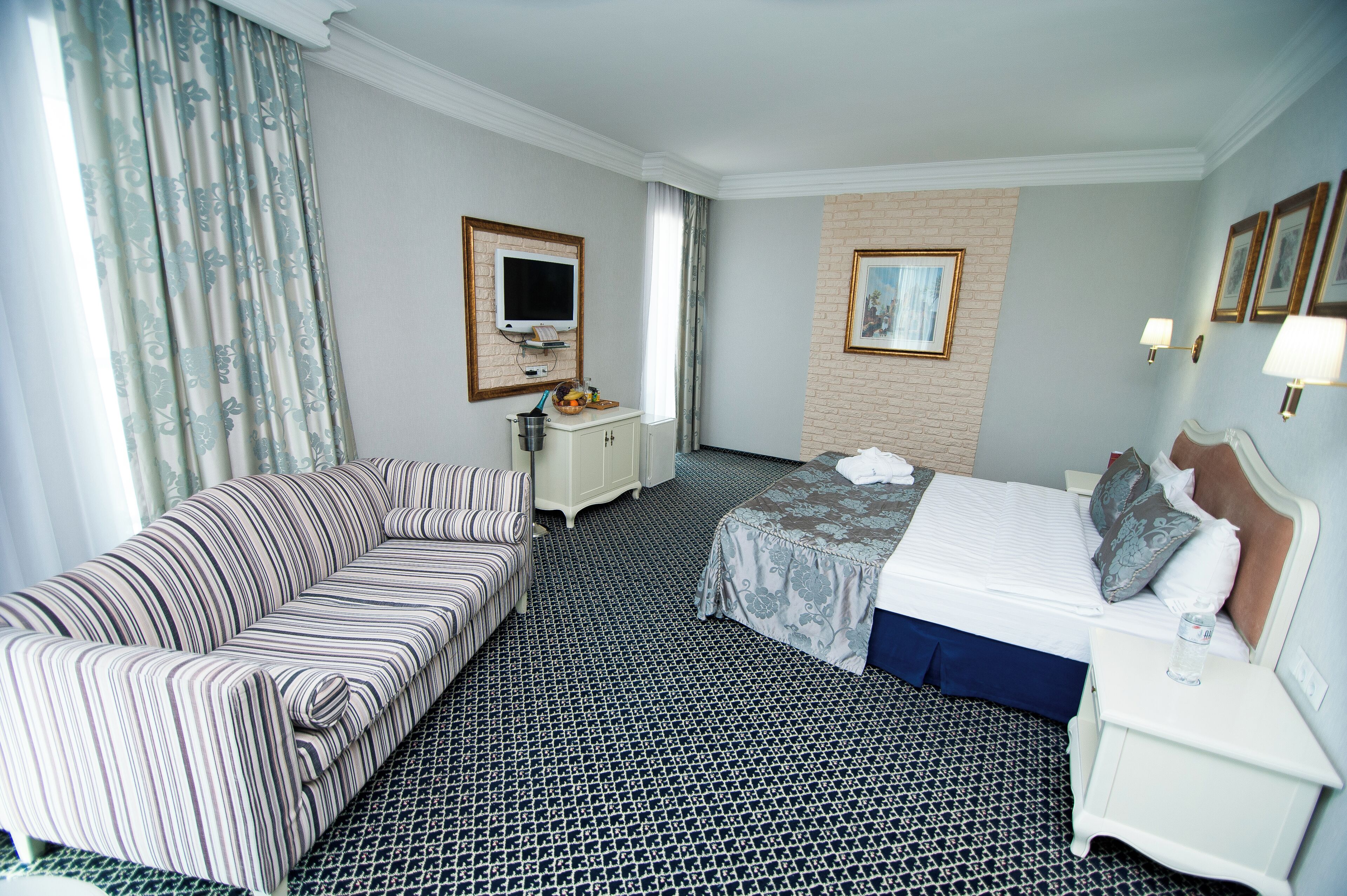 Ein Hotelzimmer mit einem gestreiften Sofa, einem Bett mit blauer Bettdecke, einem kleinen Nachttisch mit Lampe und einem an der Wand befestigten Fernseher.