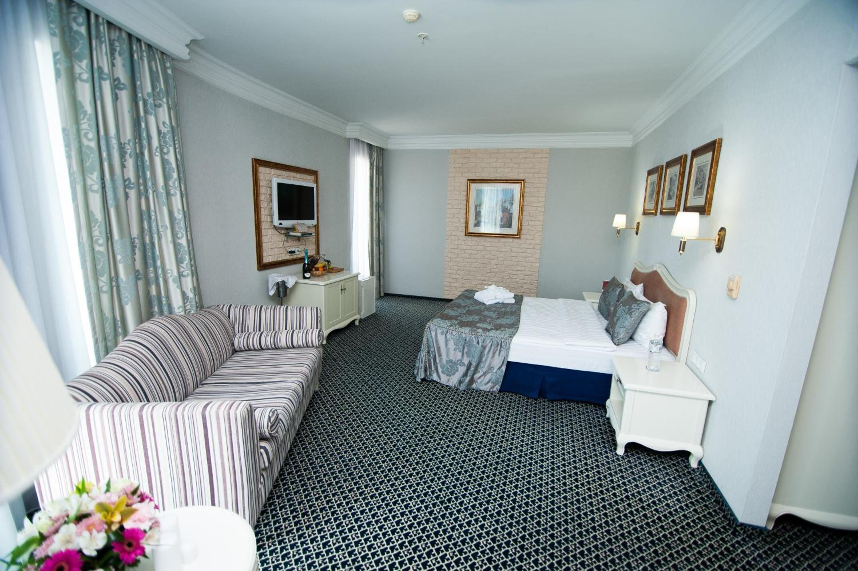 Habitación de hotel con un sofá a rayas, una cama con colcha azul, una mesita de noche pequeña, una mesita auxiliar, un televisor y un cuadro colgado en la pared.