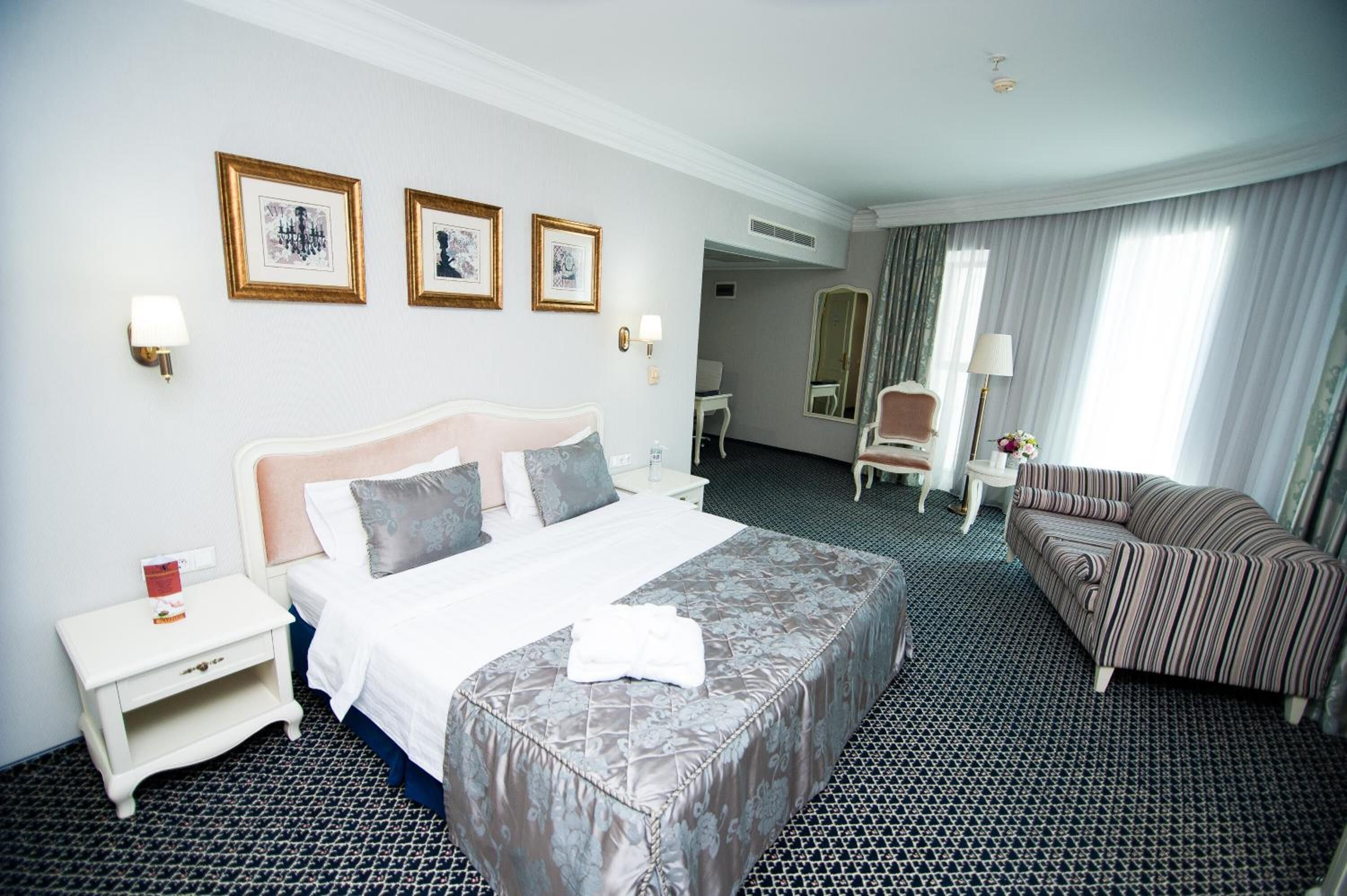 A hotel room with a large bed, a sofa, a desk, and a chair. There are three framed pictures on the wall and a mirror.