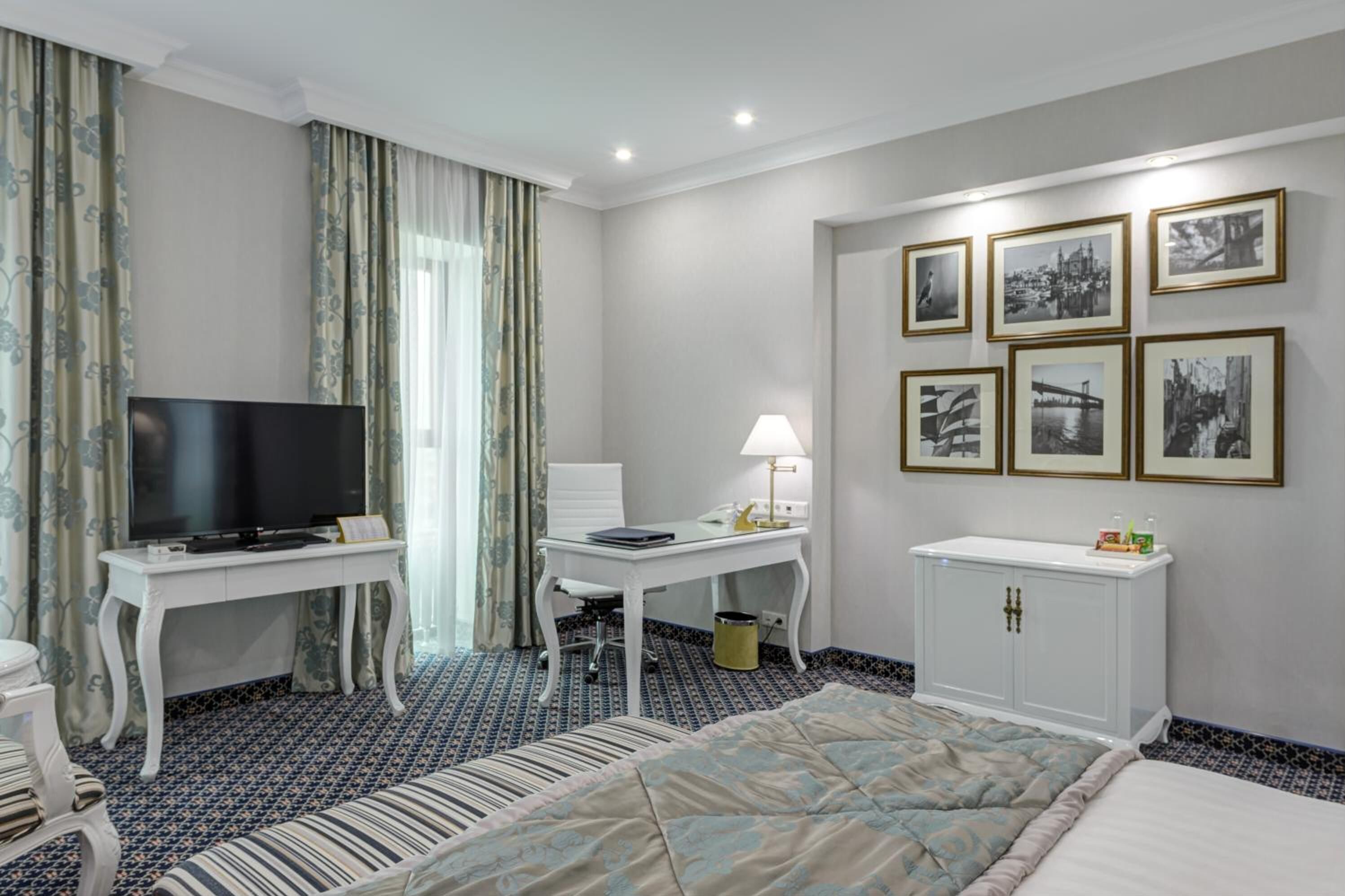 A hotel room with a bed, a TV, a desk with a lamp, a chair, and a cabinet. There are framed pictures on the wall.