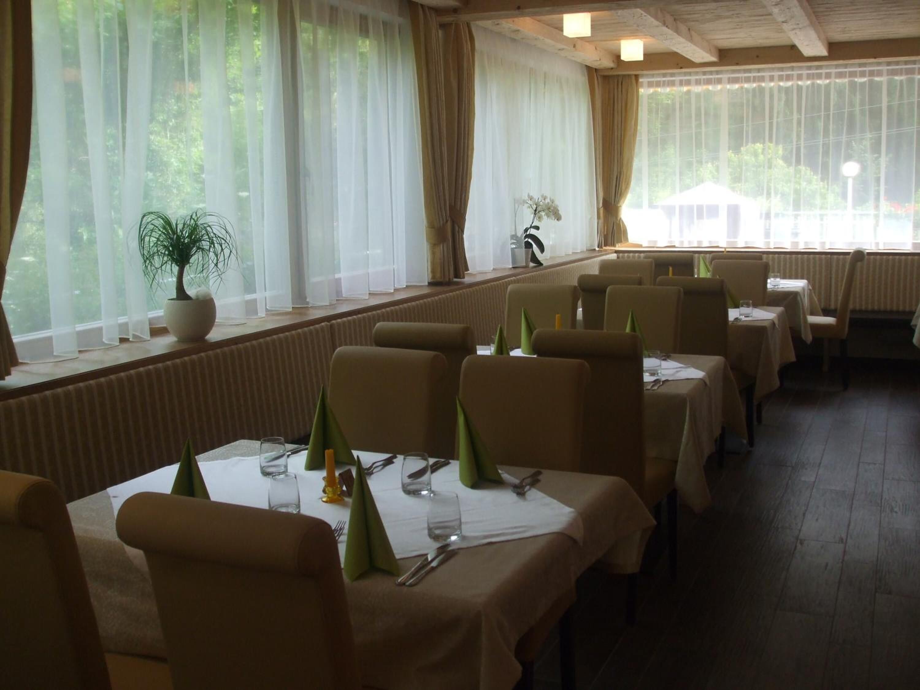 A restaurant with tables set for dining, large windows with sheer curtains, and a potted plant on the windowsill.