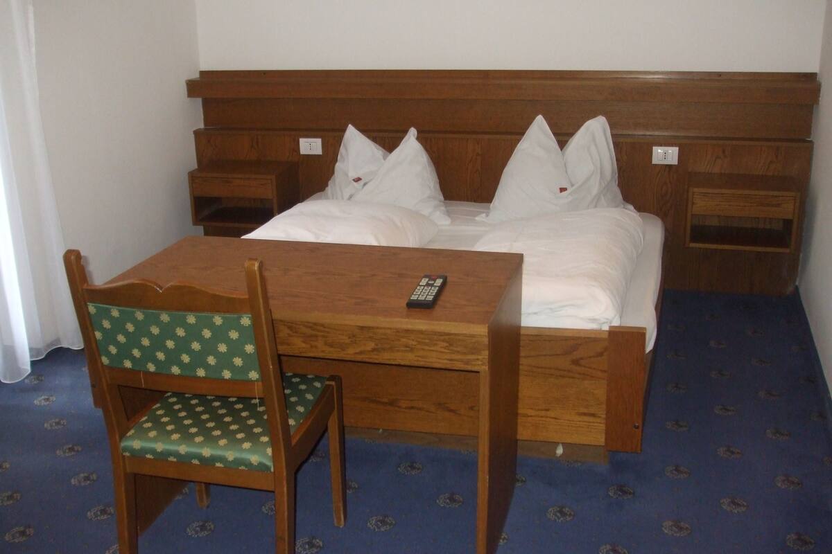 Ein Einzelbett mit Holz-Kopfteil, ein Nachttisch, ein Stuhl mit Blumenkissen und eine Fernbedienung auf dem Tisch.