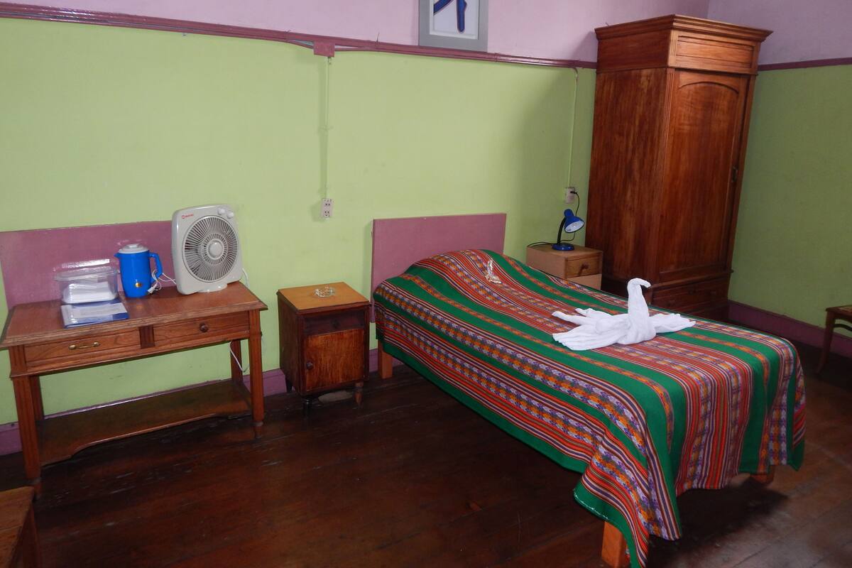 A room with a bed, a wooden desk, a fan, a lamp, and a wardrobe.