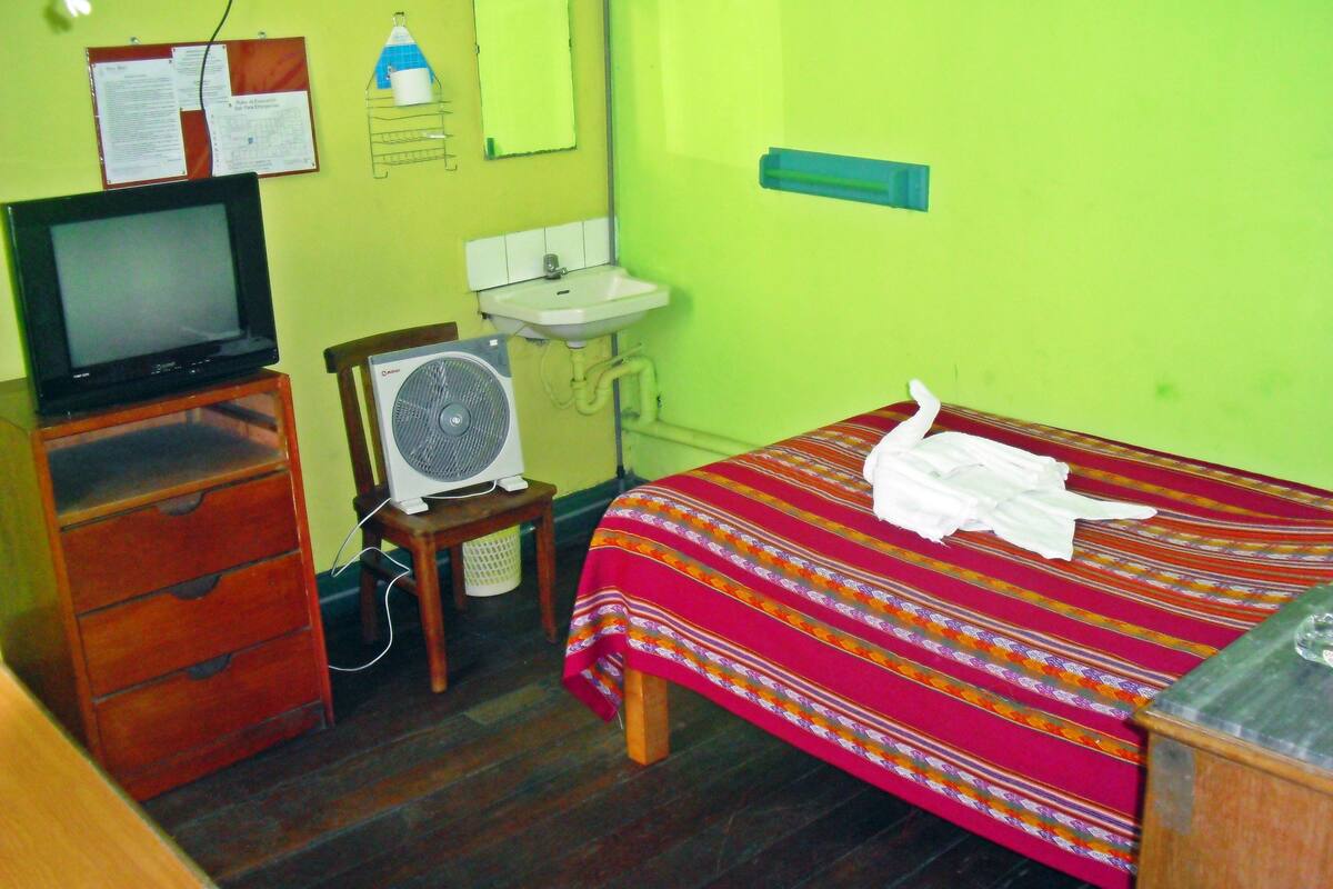 A small, simple room with a bed, a wooden dresser, a television, a fan, a sink, and a mirror.