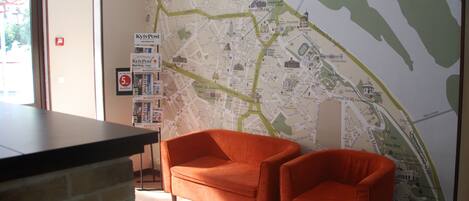 Un vestíbulo de hotel con un mapa grande en la pared, dos sillones naranjas, un mostrador de recepción y una pared de ladrillo.