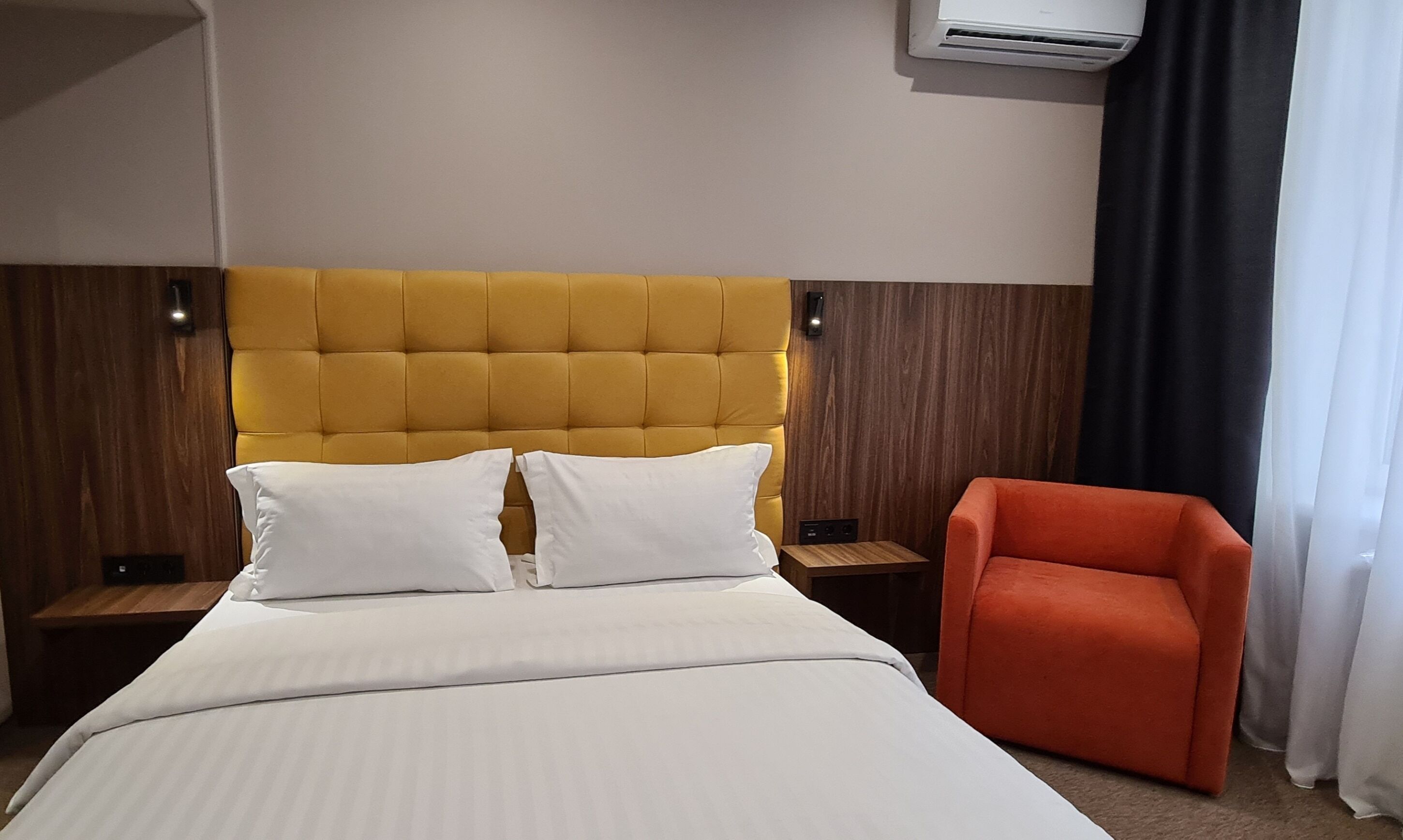 A hotel room with a large bed, two white pillows, a yellow tufted headboard, an orange armchair, a wooden nightstand, and an air conditioning unit on the wall.