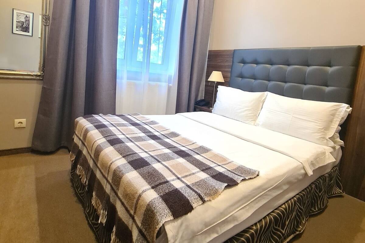 Una habitación de hotel con cama, mesita de noche, lámpara, ventana con cortinas y aire acondicionado.
