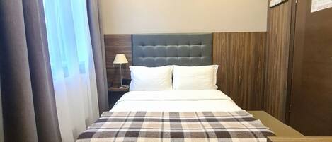 A hotel room with a bed, a nightstand, an air conditioning unit, a wardrobe, and a window with curtains.