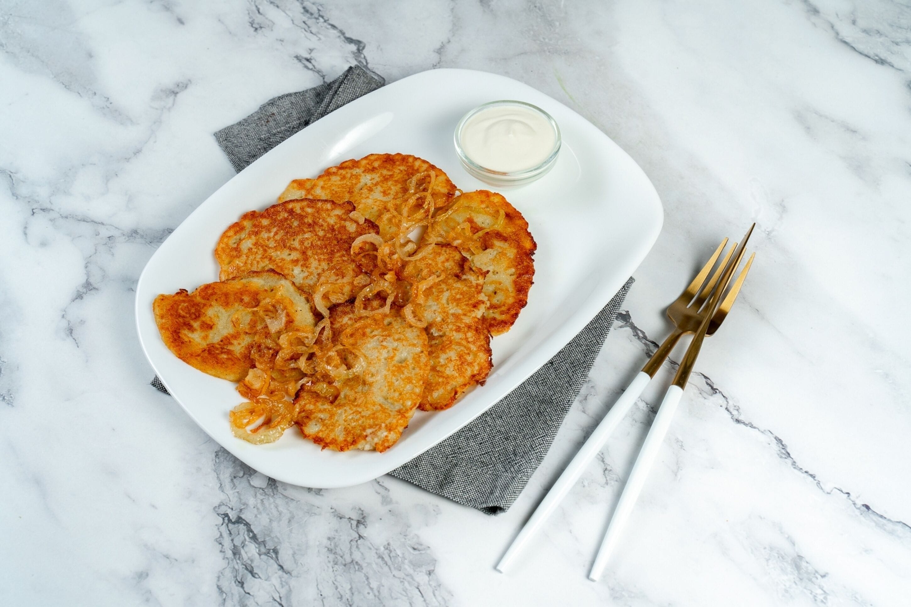 A white plate with golden-brown potato pancakes and a small bowl of white sauce, placed on a marble surface with a gray cloth napkin and gold-handled forks.