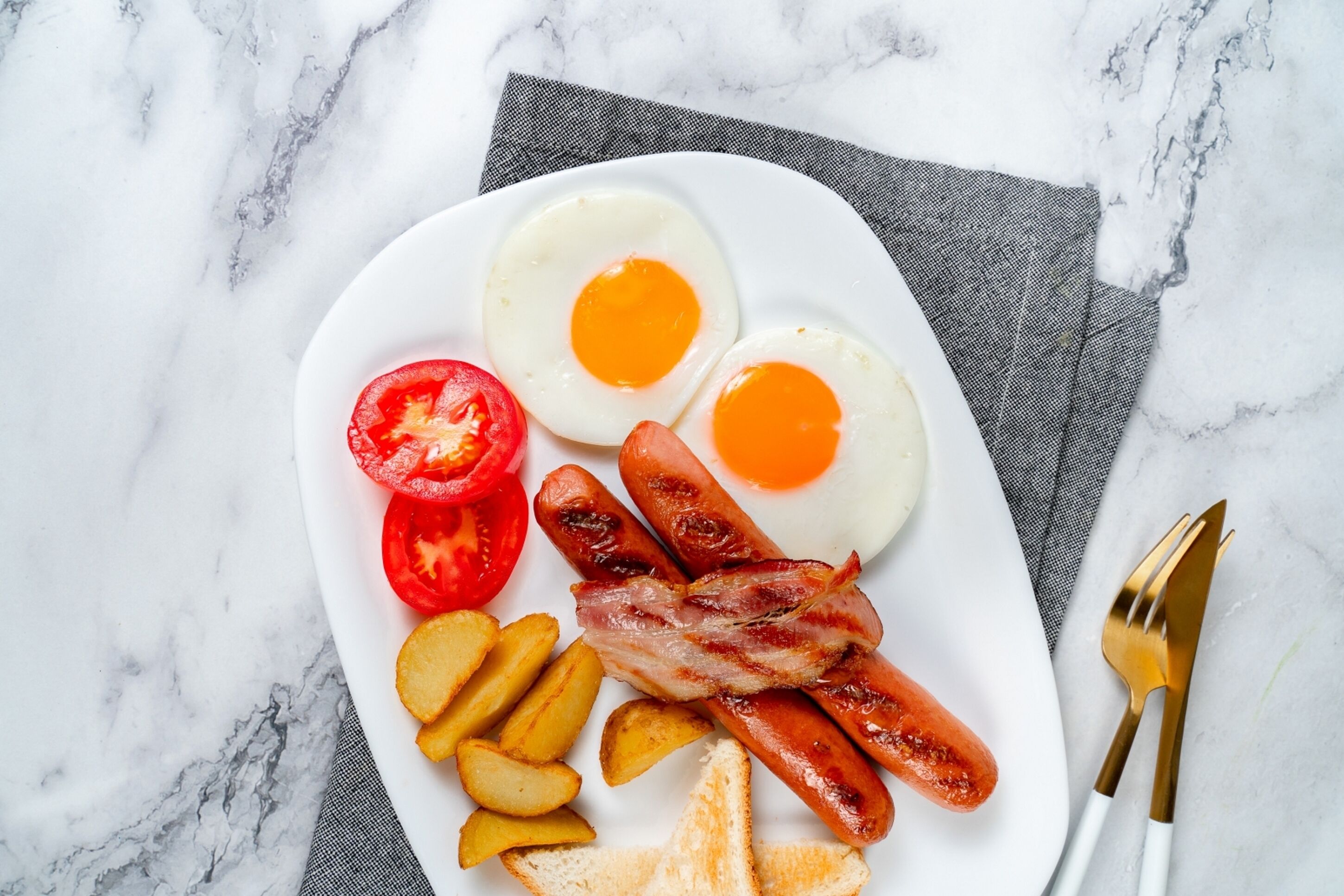 Un plato de comida para el desayuno con huevos, salchichas, tocino, tomates y tostadas sobre una superficie de mármol.