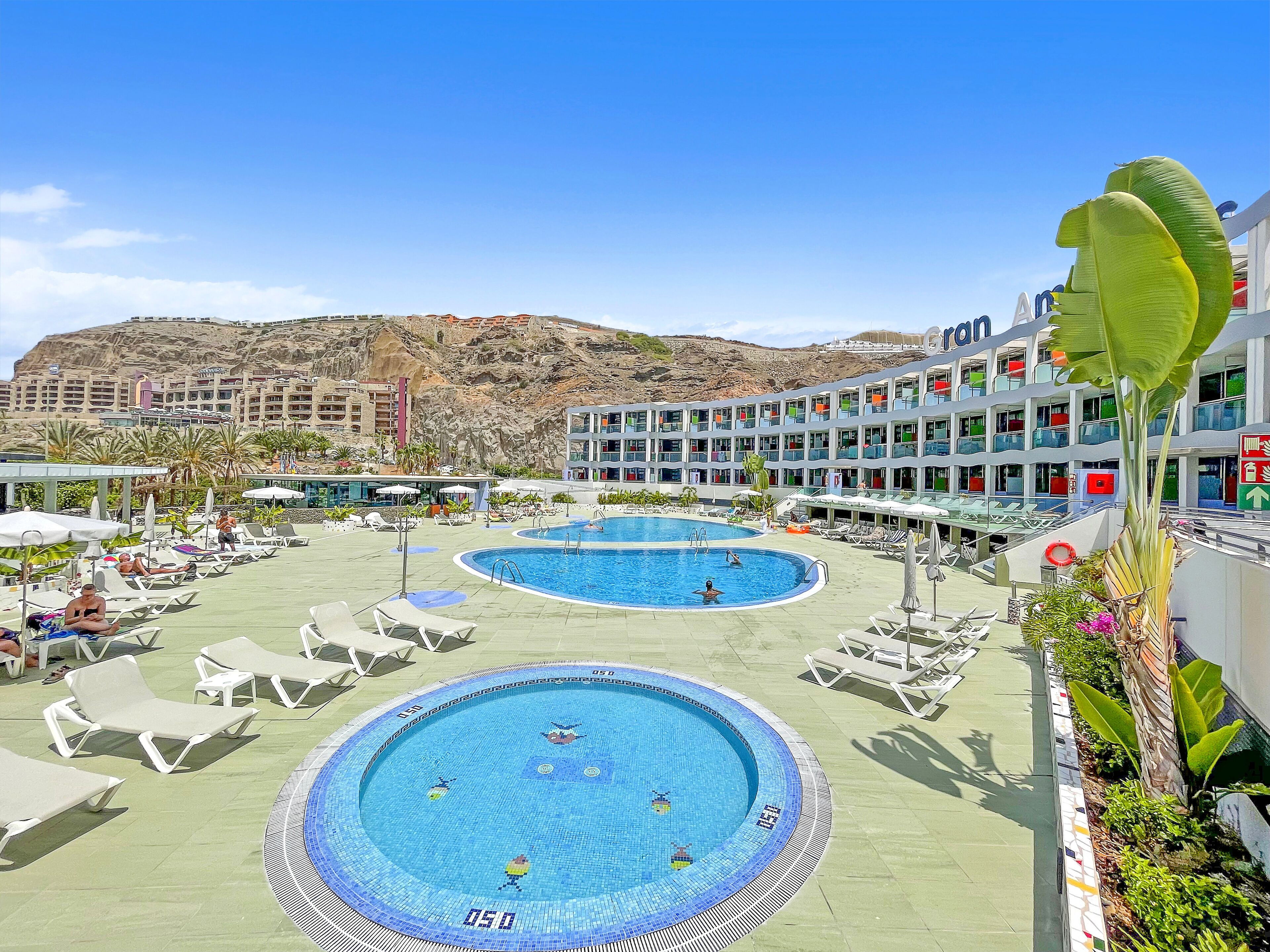 A resort with two swimming pools, sun loungers, and a view of a rocky hillside.