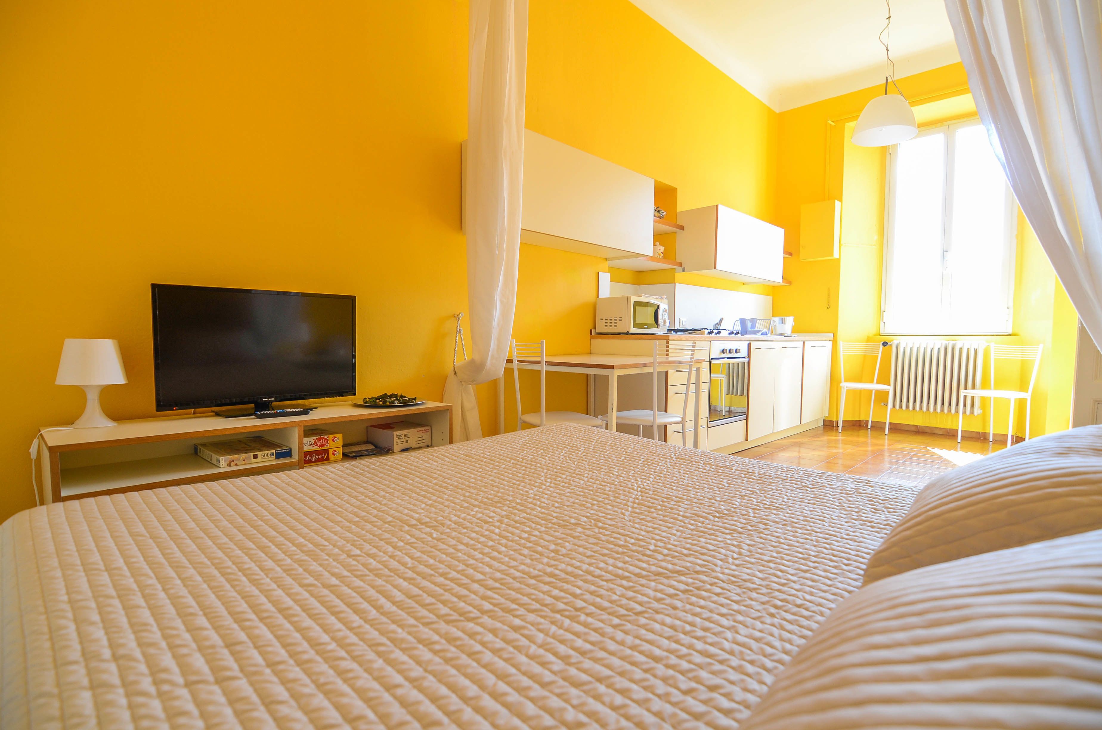 Una habitación luminosa de color amarillo con una cama, un televisor, un escritorio y una silla.
