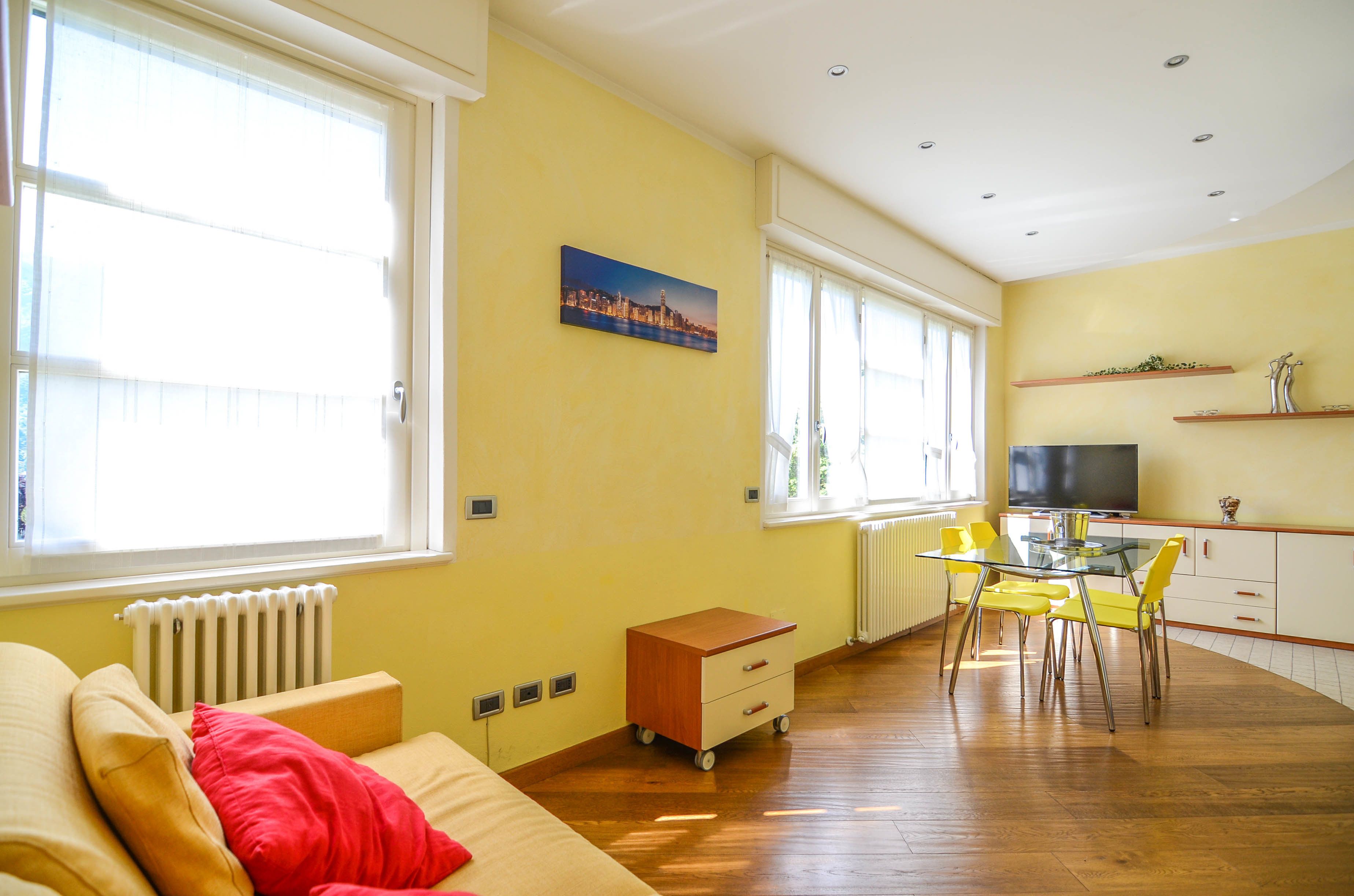 A bright living room with a sofa, a small wooden cabinet, a glass dining table with yellow chairs, and a television on a cabinet.
