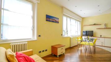 A bright living room with a sofa, a small wooden cabinet, a glass dining table with yellow chairs, and a television on a cabinet.