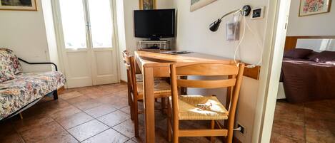 A room with a wooden dining table, chairs, a television, and a bed with a brown blanket.