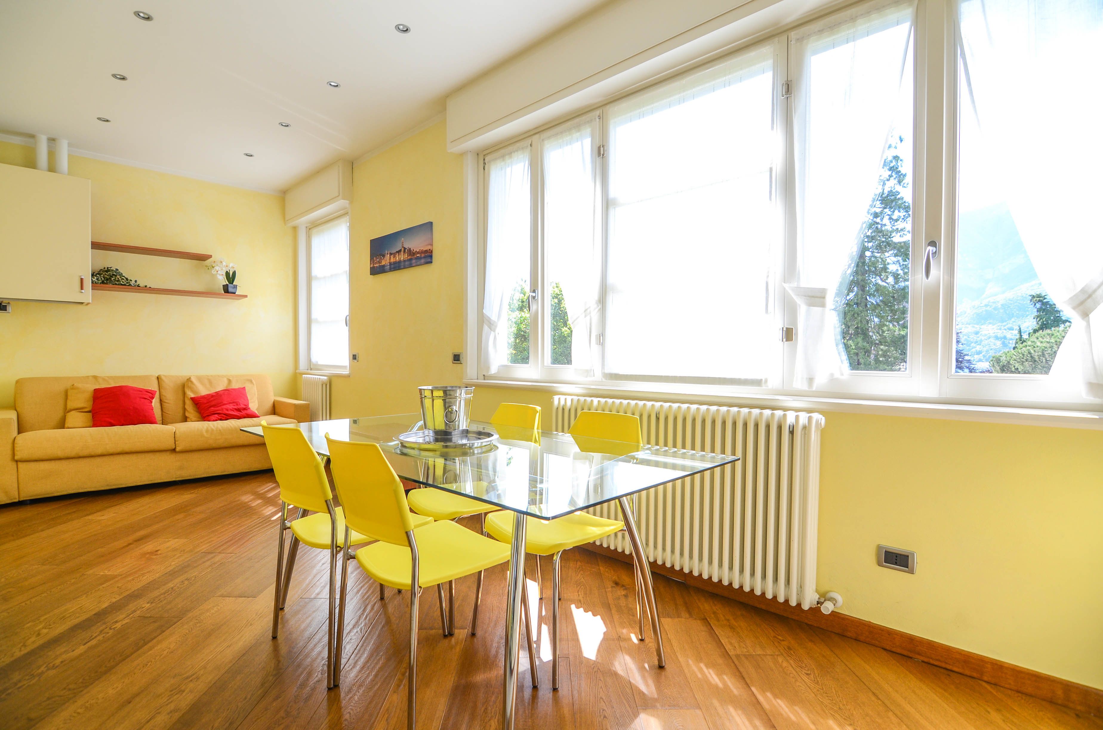 Une pièce lumineuse avec une table en verre, des chaises jaunes, un canapé beige agrémenté de coussins rouges et une grande fenêtre donnant sur un paysage verdoyant.