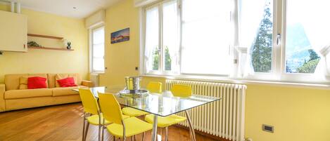 Une pièce lumineuse avec une table en verre, des chaises jaunes, un canapé beige agrémenté de coussins rouges et une grande fenêtre donnant sur un paysage verdoyant.