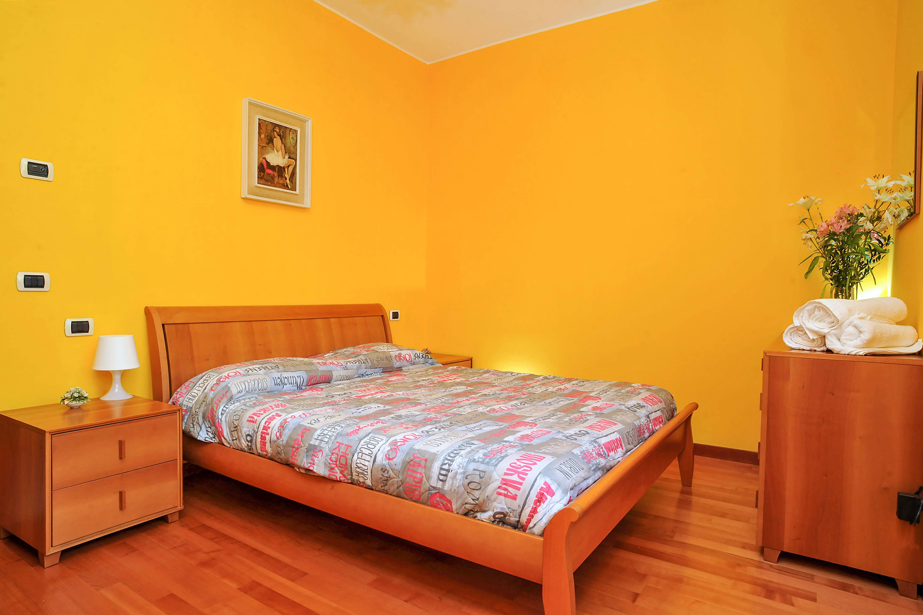 A bedroom with a wooden bed, a nightstand with a lamp, a dresser, and a framed picture on the wall.