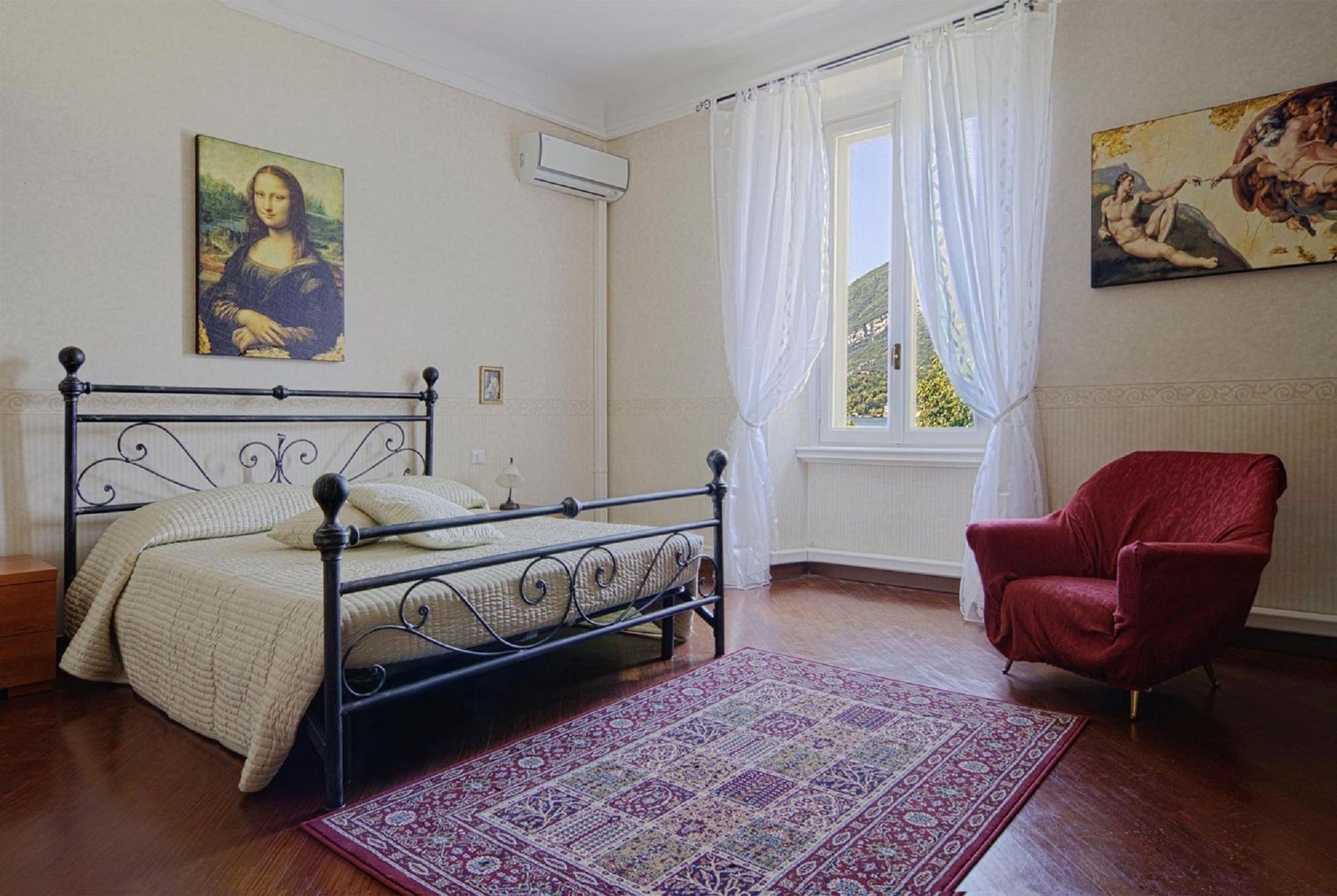 A bedroom with a bed, a red armchair, a large window with curtains, and two paintings on the walls.