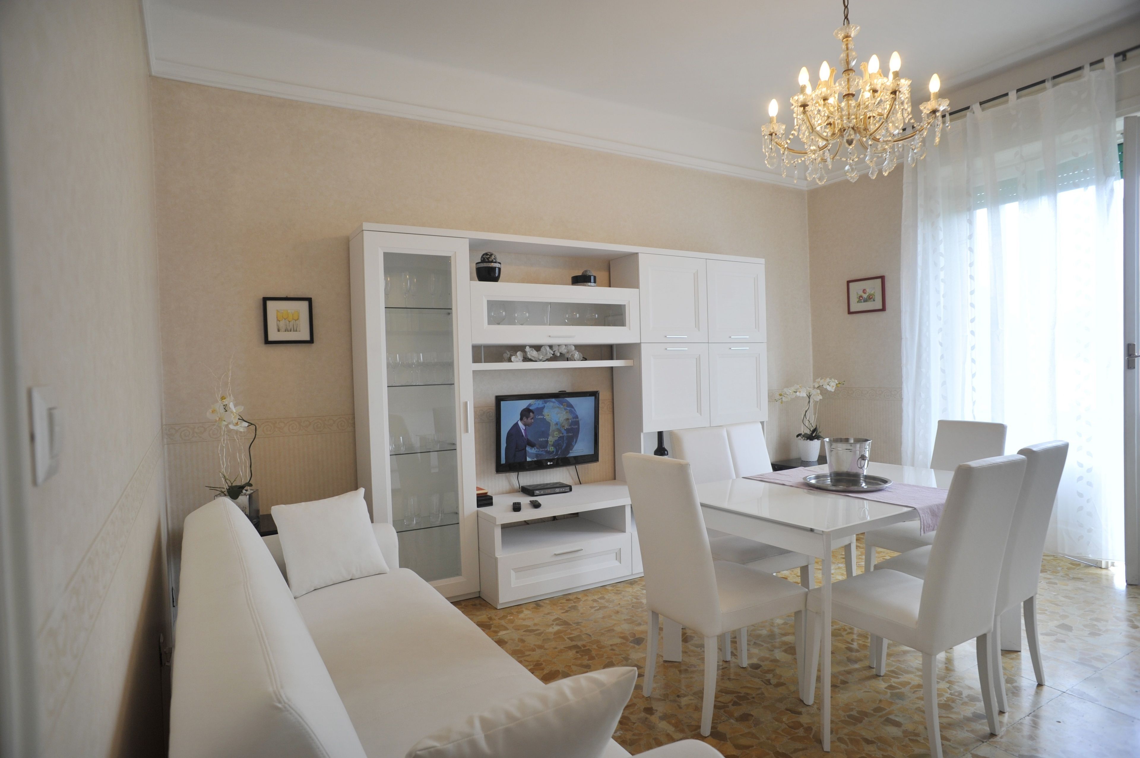 Ein modernes Wohnzimmer mit einem weißen Sofa, einem Esstisch mit Stühlen, einem Fernseher und einer Kristallleuchter.