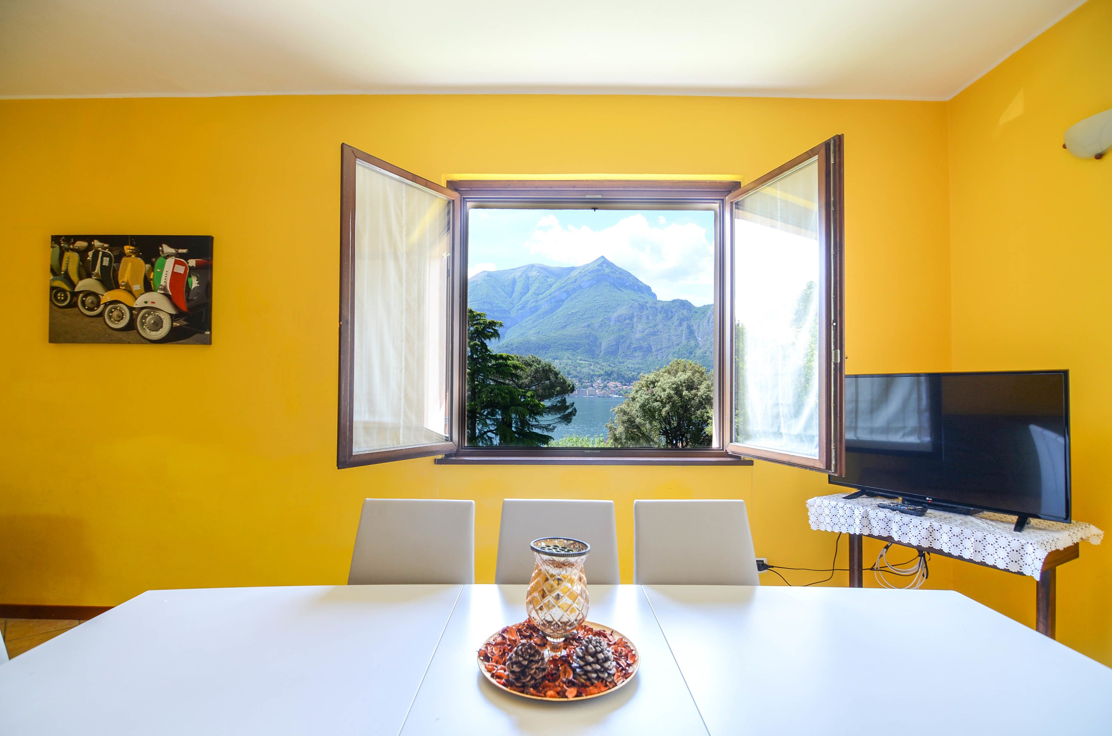 A dining area with a table, chairs, a television on a stand, a window with an open view of mountains, and a painting on the wall.
