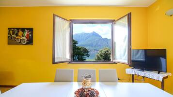 A dining area with a table, chairs, a television on a stand, a window with an open view of mountains, and a painting on the wall.