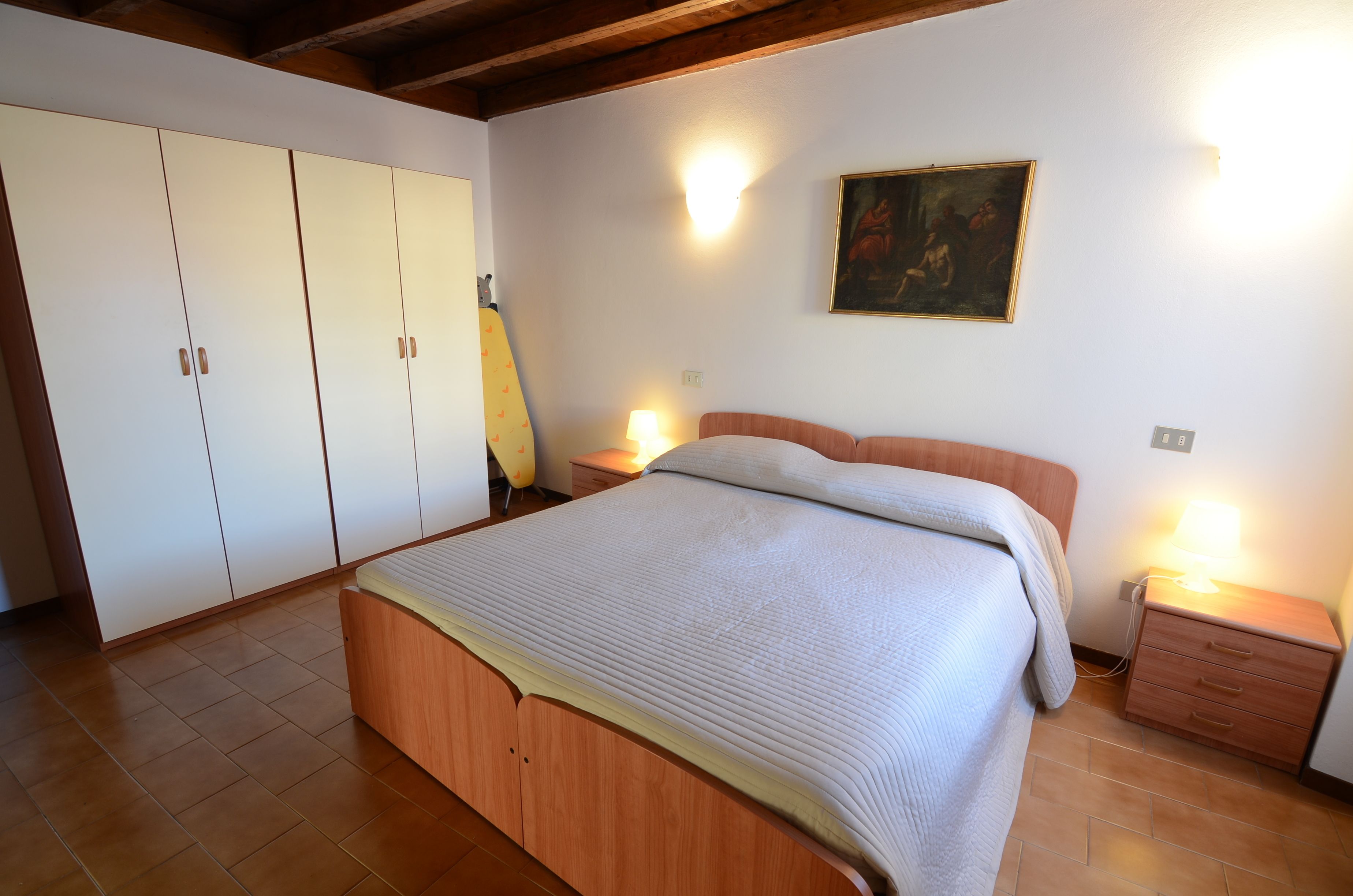 A bedroom with a wooden bed, a nightstand with a lamp, a wardrobe, and a painting on the wall.
