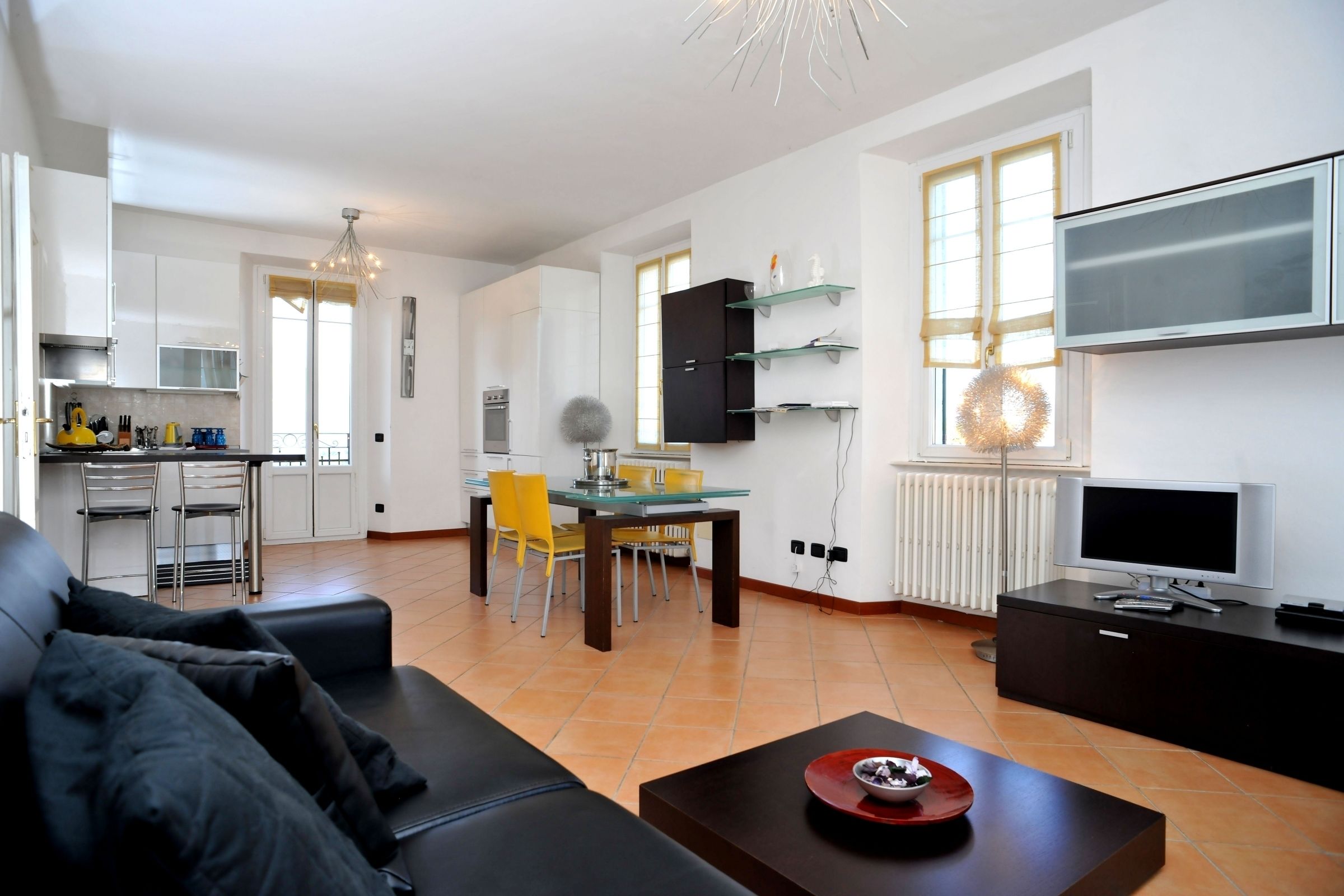 A modern living room with a sofa, a coffee table, a dining area with chairs, a kitchen with cabinets, and a TV mounted on the wall.